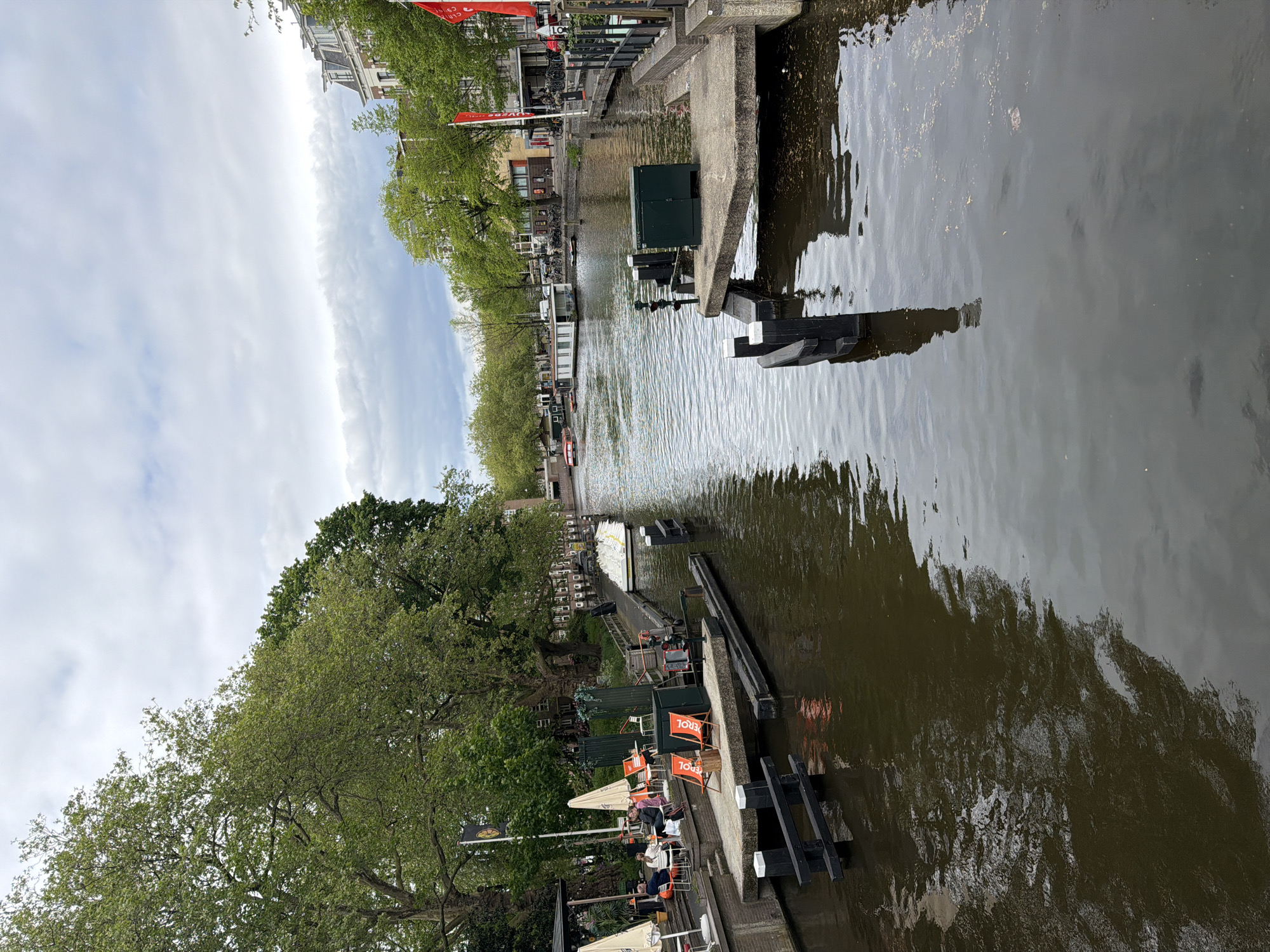 The image shows a canal in Amsterdam, Netherlands. The view is oriented along the canal, with water filling most of the right half of the frame. Along the left bank of the canal, there is a tree-lined walkway with buildings and shops visible in the background. People are sitting at outdoor cafes and patios alongside the canal. The canal itself has various concrete and metal structures along the edges, possibly for boat mooring or as part of the canal's infrastructure. The sky is overcast with scattered clouds.