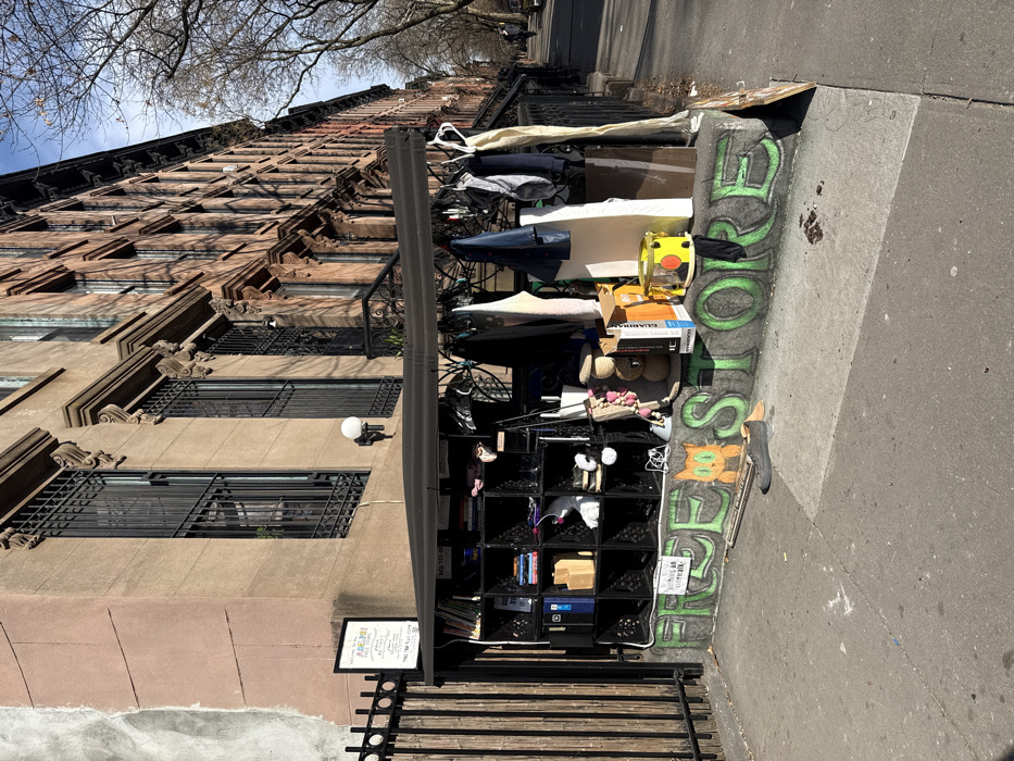 The image depicts an outdoor community "free store" situated on Adelphi Street in Brooklyn, New York City, United States of America. The scene is captured on a clear, sunny day, with a bright blue sky visible above a row of residential brownstone buildings.
The free store occupies the sidewalk level of a stoop, directly in front of a three-story brownstone building. A black canopy shelters the setup. The store's structure consists of black milk crates stacked to form shelves, displaying various items including books, small boxes, footwear, and miscellaneous objects. Clothing, such as shirts and jackets, hangs from a rod underneath the canopy. The front edge of the stoop is marked with green graffiti spelling "FREE STORE," featuring a cartoon cat face. A small white sign reading "nothing on sidewalk" is visible within the graffiti. On the stoop, beside the crates, additional items include cardboard boxes, a yellow cylindrical container, a stack of wooden rings, and a white rectangular object. A single dark shoe and a rolled object lie on the concrete sidewalk in front of the stoop.
A black metal fence runs along the left side of the scene, attached to the building. A white, framed sign hanging on this fence identifies the location as the "ADELPHI STREET FREE STORE." The adjacent brownstone buildings feature ornate cornices and black wrought iron security grates on their windows. A white spherical wall light is mounted on the building above the free store. Bare trees line the sidewalk down the street to the right, consistent with winter or early spring. No individuals are prominently visible in the immediate foreground or midground; however, faint figures can be seen walking in the distance down the street on the right.