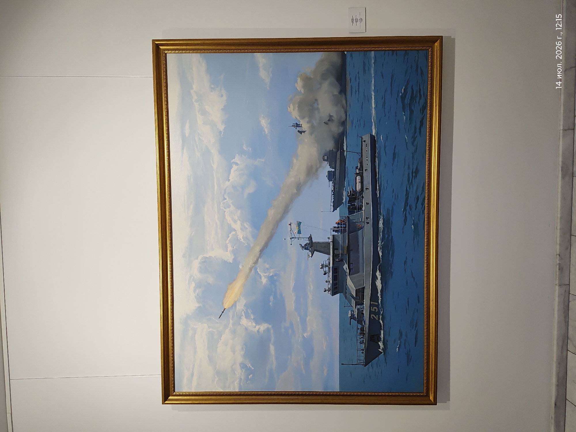 An oil painting in a golden frame depicts a naval scene. In the foreground, a grey military vessel with the number "251" on its side cuts through choppy blue water, leaving a white wake. A missile is seen launching from behind the vessel, leaving a trail of smoke against a cloudy sky. In the background, another vessel is partially obscured by a large plume of smoke. A small white label is affixed to the wall to the right of the painting, and a timestamp "14 июл. 2026 г., 12:15" is visible in the lower right corner. The painting is displayed on a white wall in what appears to be an art gallery or museum, with a marble baseboard along the bottom of the wall. The location is stated to be in the city of Astana, Kazakhstan.