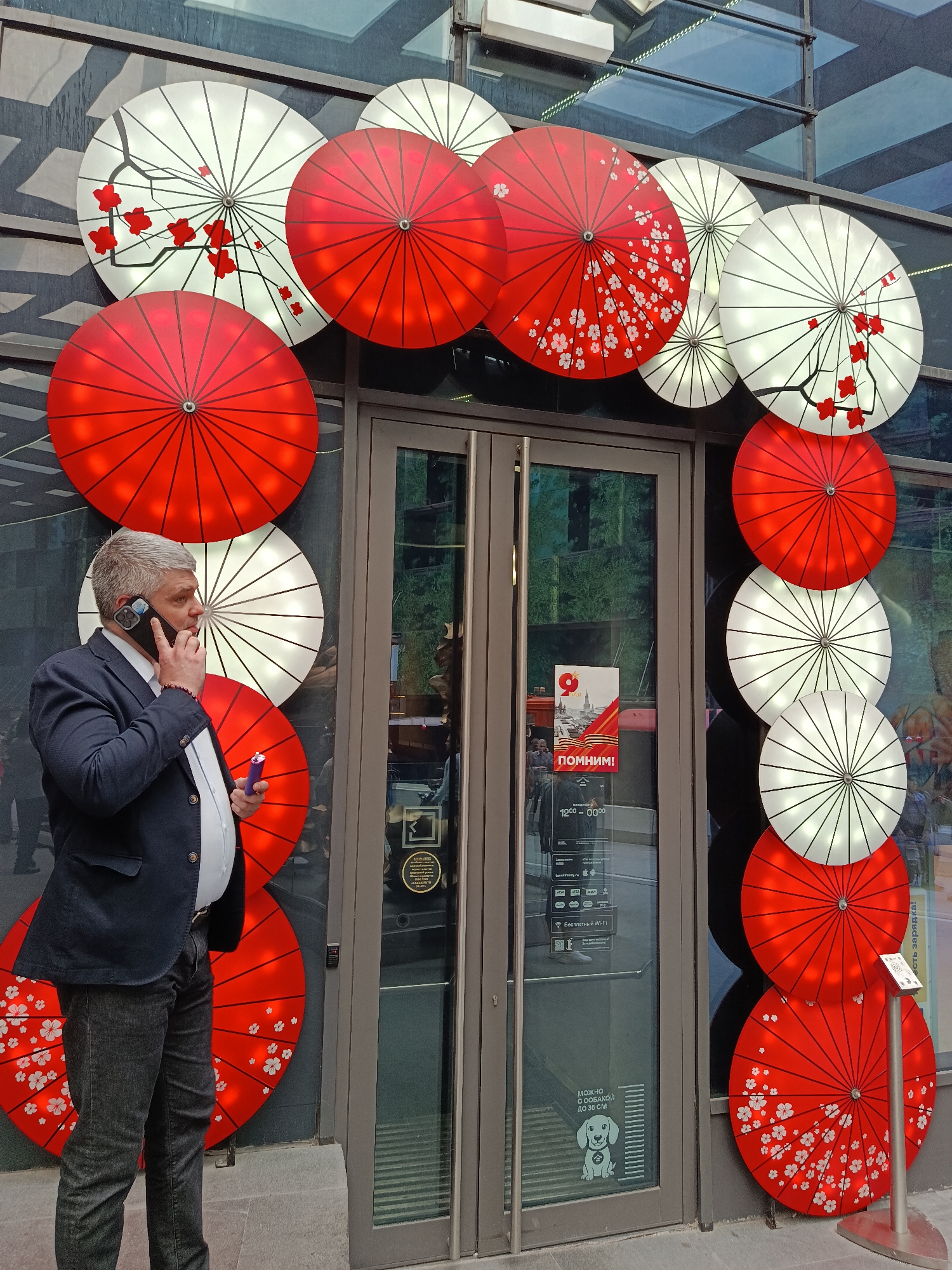 The image shows a middle-aged man in Moscow, Russia, standing in front of a building. The building has glass doors flanked by large, decorative umbrella-shaped lights, alternating between red and white. Some of the umbrellas are decorated with floral patterns. The man is wearing a dark blazer, white shirt, and dark jeans. He is holding a purple cylindrical object in one hand and talking on a mobile phone held to his ear. A sign on the glass door has text in Russian, including the word "ПОМНИМ!" (We Remember!), opening hours, and a graphic of a dog with accompanying text indicating dogs under 35 cm are permitted. A metal sign on a stand is visible to the right of the door. The building's facade has a grid-like pattern.