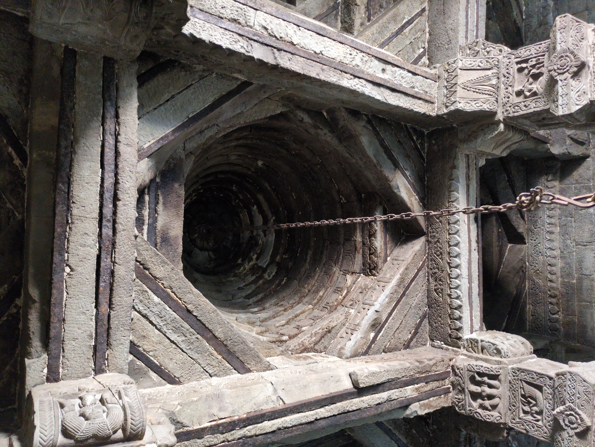 This image depicts the interior of an ancient stone structure, likely a temple or a well. The perspective is looking upwards towards a circular, tiered opening at the apex of the structure. Thick, rough-hewn stone beams crisscross overhead, supporting the ceiling. Some of these beams appear to be reinforced with metal bars, suggesting a blend of ancient and possibly later construction.
Suspended from the center of the opening is a heavy, dark metal chain. The stone elements of the structure are intricately carved, particularly at the base of the supporting columns where figures and decorative motifs are visible. These carvings suggest a rich artistic and cultural heritage. The stone itself appears weathered and aged, with a rough texture and visible signs of erosion. The lighting is dim, with shadows creating a sense of depth and mystery, particularly within the dark, circular opening above. The overall impression is one of historical significance and architectural complexity. The location is unknown.