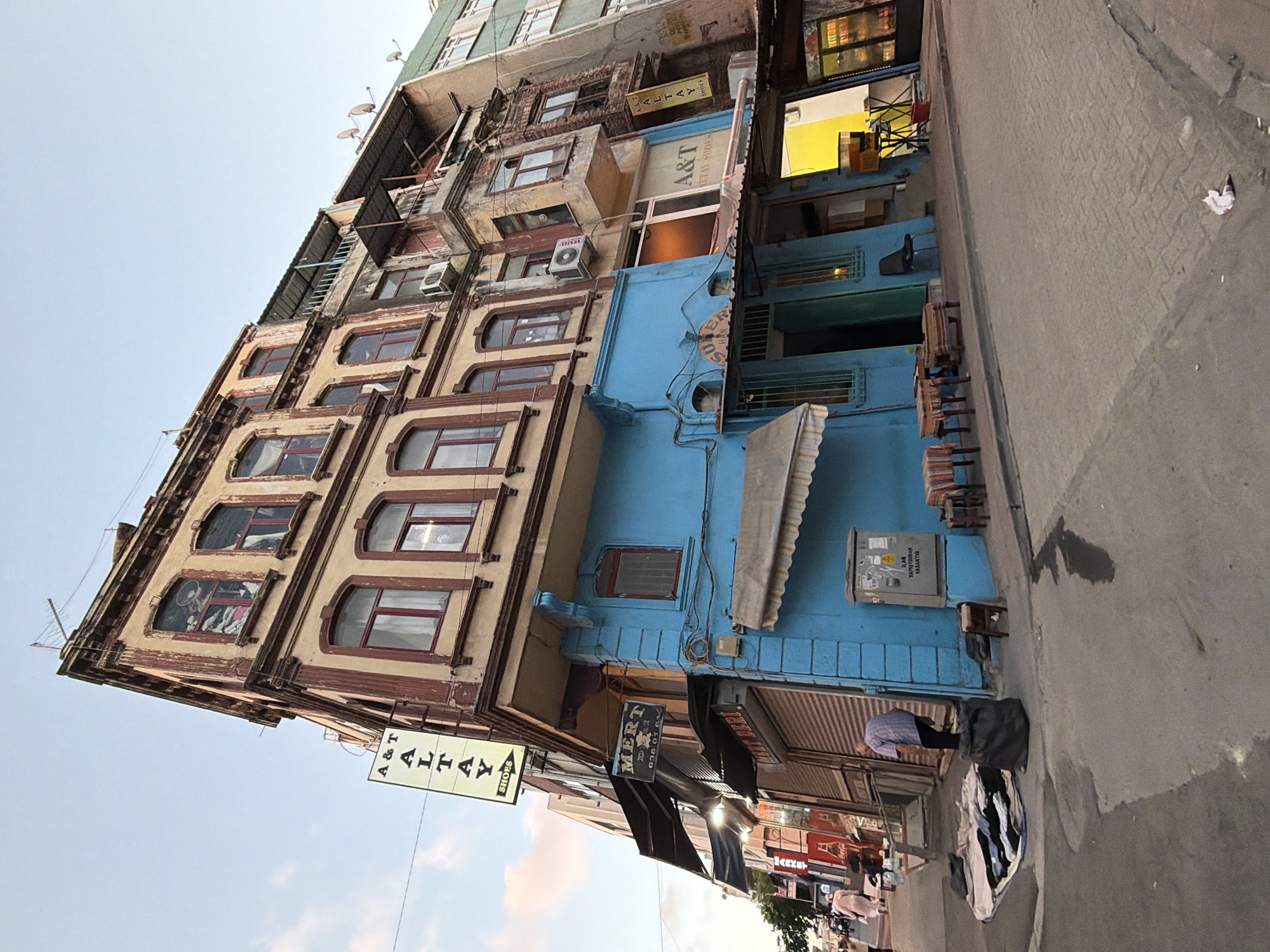 This is a vertical, eye-level, full shot of a narrow, bustling street scene in Istanbul, Turkey, dominated by the facades of old buildings. The buildings are a mix of colors and architectural styles, with some appearing weathered and others more recently painted. The sky is a pale, overcast blue.
The primary subject is the street itself, lined with shops and businesses. On the left, a multi-story building with tan and brown facades features numerous windows, some with dark red frames, and ornate detailing. Signs are visible, including one that reads "A & T ALTAY SHOES". Further down, a vibrant blue building interrupts the streetscape, with a light blue awning and several windows. Adjacent to this blue building, another shop sign reads "MERT DERI". The street level appears to be lined with storefronts, some with metal shutters.
Objects in the scene include numerous chairs, some stacked and others arranged near storefronts, suggesting outdoor seating or displays. Trash and debris are scattered on the sidewalk and street. Electrical boxes and wiring are also visible, indicative of the urban environment.
The activity appears to be daytime commerce. While few people are clearly visible, there are indications of pedestrian traffic and shop activity. One person in the foreground, wearing a light-colored shirt and dark pants, is bent over a pile of clothing or fabric on the ground. Another person, in the background on the right, wears a light pink garment.
Notable details include the weathered appearance of some buildings, suggesting age and history. The mix of signs in Turkish and English hints at a tourist-friendly area. The overall impression is of a lively, somewhat cluttered, and well-worn urban environment. The time of day appears to be late afternoon or early evening, given the lighting conditions.
