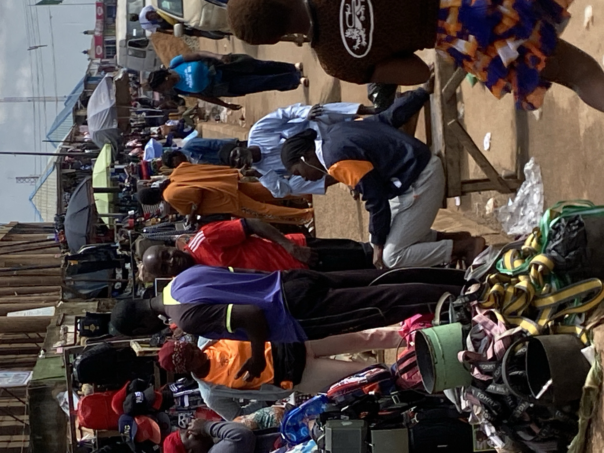 The image depicts a bustling market scene in Bukuru, Nigeria. In the foreground, several individuals are engaged in various activities. A woman wearing a blue shirt and dark pants walks away from the viewer, her back to the camera. To her left, a man in a light blue traditional outfit squats, looking to his right. Another individual, seen from behind, sits on a wooden bench, wearing a dark blue and orange jacket and grey pants. To the left, a pile of bags and buckets are displayed, some with colorful straps.
Behind the foreground figures, the market extends further. Stalls are set up with merchandise, including clothing and accessories, many of which are displayed on racks or tables. Umbrellas and awnings provide shade from the sun. People are seen walking through the market, browsing, and interacting. In the background, buildings with corrugated roofs are visible, and a white van is parked on the right side of the frame. The ground is a dusty, reddish-brown dirt path, typical of many Nigerian marketplaces. The overall impression is one of a lively and active commercial environment.