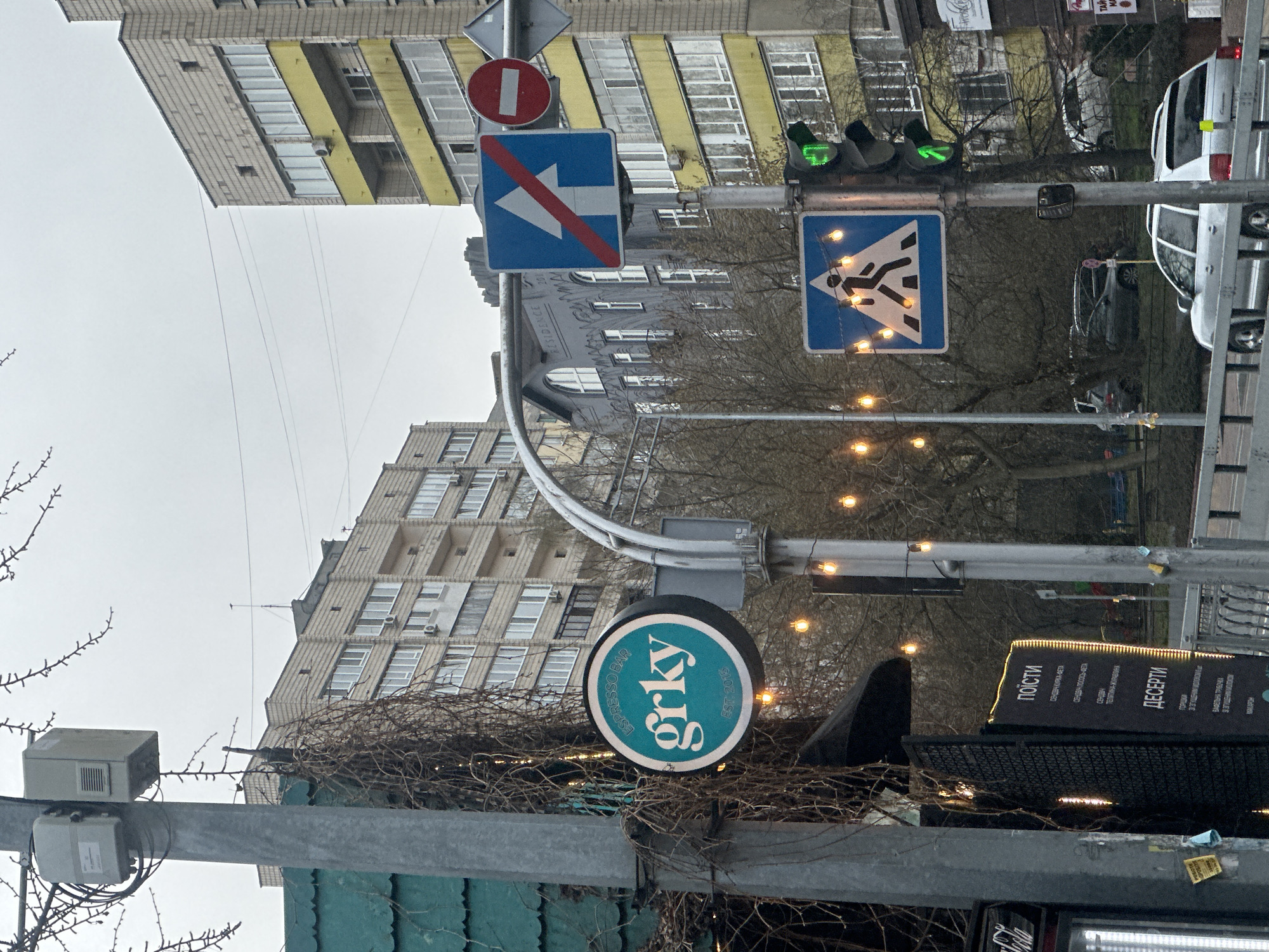The image, taken in Kyiv, Ukraine, shows a street scene with an "grky Espresso Bar" sign attached to a metal pole. Above the sign are various traffic signs including a "no entry" sign (red circle with a white horizontal bar), a "end of lane" sign (blue square with a white arrow crossed out by a red stripe), and a pedestrian crossing sign (blue triangle with a figure crossing a street). The traffic light is showing a green signal for pedestrians. Buildings with windows are visible in the background. A menu board with lettering that reads "поїсти" and "ДЕСЕРТИ" is also present. Wires and branches intertwine around the signage and pole. A Coca-Cola sign is partially visible at the bottom of the frame.