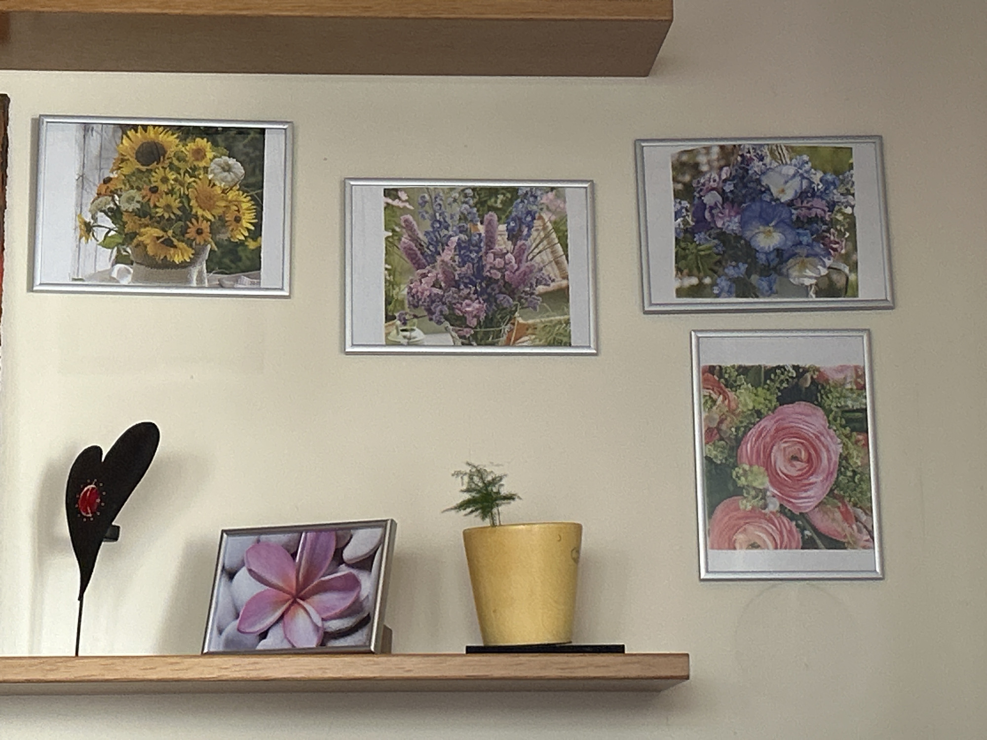 The image shows a shelf with several framed pictures of flowers and a decorative heart-shaped object. On the top shelf, there are three framed pictures. The leftmost picture displays a bouquet of sunflowers and other yellow flowers in a vase. The middle picture shows a bouquet of purple and pink delphiniums and other flowers. The rightmost picture features a bouquet of blue and purple pansies and other flowers.
Below these, on a lower shelf, are more items. To the left, there is a black decorative object shaped like a heart with a red circular embellishment. Next to it is a framed picture of a pink plumeria flower resting on pebbles. In the center, a small green plant in a yellow pot sits on a black coaster. To the right, on the wall, is a framed picture of two pink ranunculus flowers surrounded by greenery. The scene appears to be indoors, possibly a home, with a light-colored wall and wooden shelves.