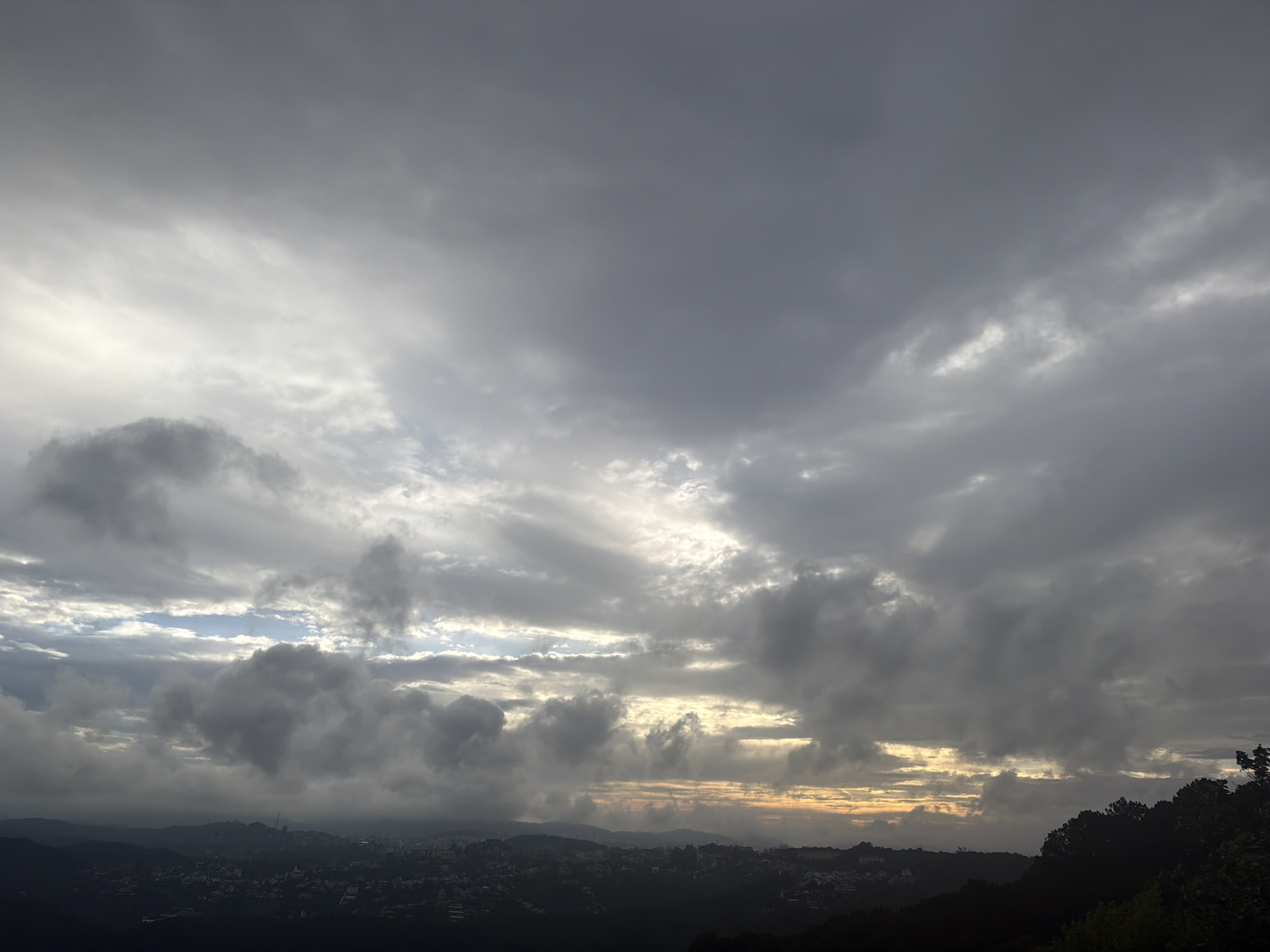 The image depicts a panoramic view of a city nestled among rolling hills under a dramatic, overcast sky. The foreground is dominated by dark, silhouetted trees on the right, framing a vast expanse of clouds. The city, appearing as a collection of small buildings and structures, is spread across the middle ground and stretches into the distance, partially obscured by atmospheric haze and the darkening landscape. The sky is filled with thick, textured clouds in various shades of gray, with some areas showing lighter patches where sunlight struggles to break through. The overall impression is that of a moody, possibly pre-dawn or post-sunset scene, with the city lights faintly visible in the distance. The environment suggests a mountainous or hilly terrain surrounding a populated area. There are no discernible people or specific actions visible in the image, only the static landscape under a dynamic sky. The location is identified as Xuân Thành, Vietnam.