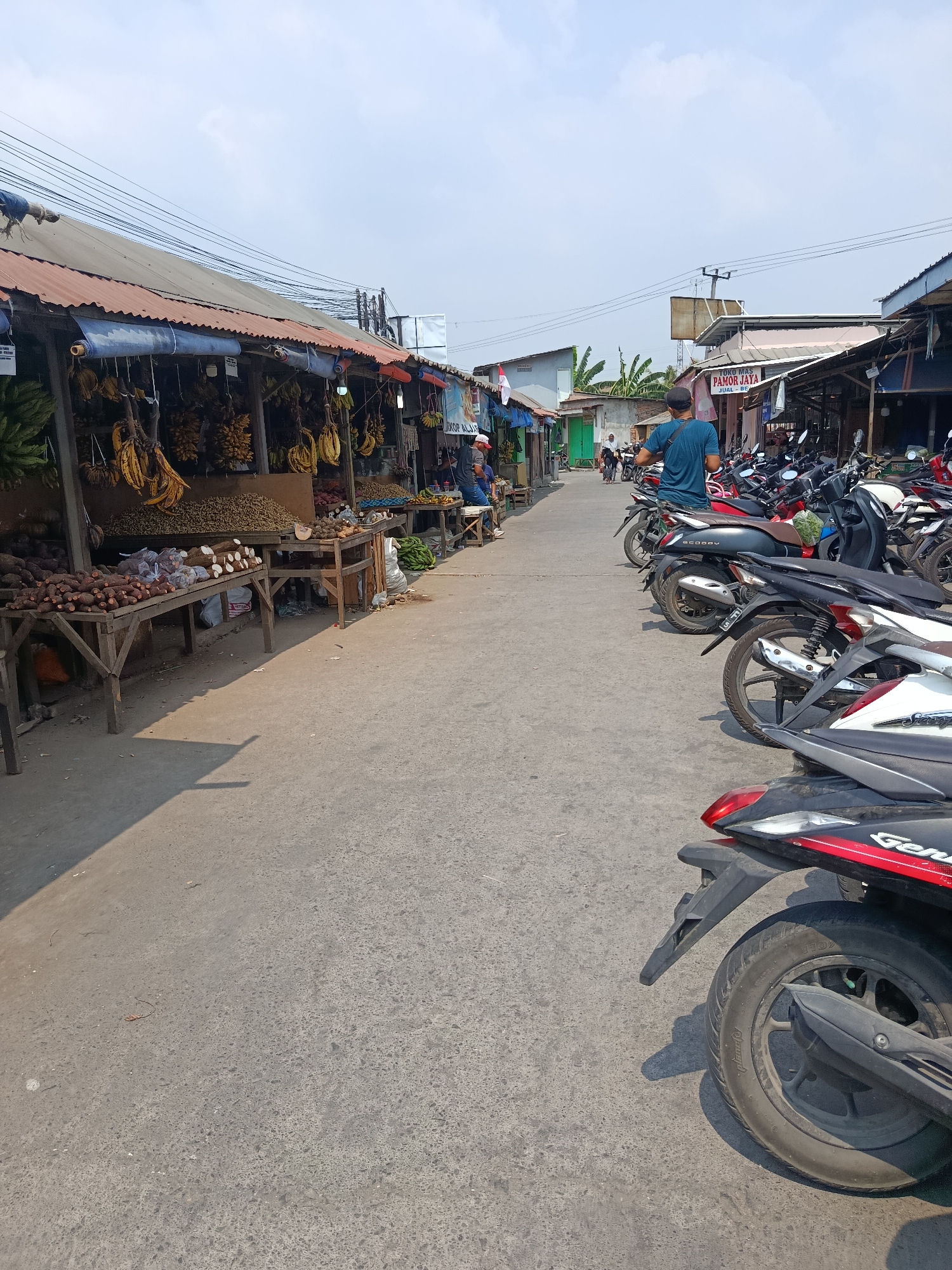 This is a vertical, eye-level, medium-shot photograph of a street market in Wanasari, Indonesia. The market is bustling with activity, and the street is lined with stalls selling various produce. In the foreground, motorcycles are parked along the right side of the street. The stalls on the left are covered with corrugated metal roofs and display bananas, yams, and other root vegetables. People are seen browsing the stalls and walking down the street. The sky is clear and blue, with a few wispy clouds. The overall atmosphere is one of a typical Indonesian street market on a sunny day.