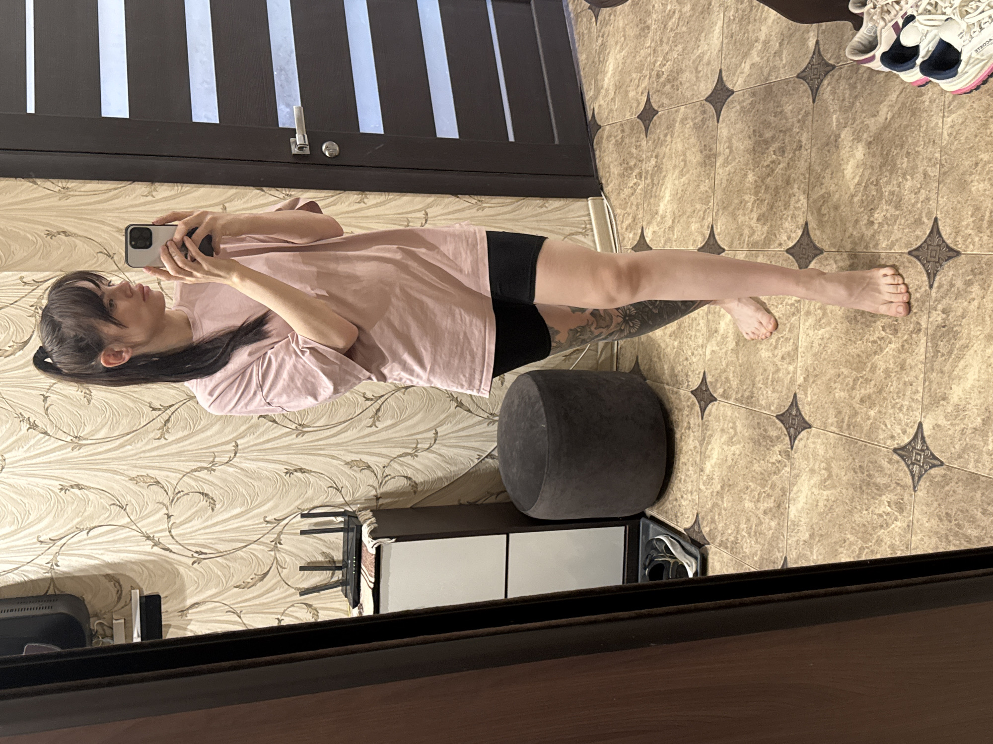 A young woman, appearing to be in her late teens or early twenties, is taking a full-body mirror selfie indoors. She has dark hair styled with bangs and tied back in a ponytail, and she wears an oversized light pink t-shirt and black shorts. A large, intricate tattoo, possibly floral or nature-themed, is visible on her left thigh. She is barefoot and holds a light-colored smartphone with multiple cameras in both hands, looking directly at the mirror. Her expression is neutral.
The setting appears to be a residential room. The background features a dark brown door with three vertical white panels. The wall behind the woman is covered in light-colored, possibly beige, wallpaper adorned with an elegant, repeating vine or floral pattern in a slightly darker shade. The floor is tiled with light brown/beige square tiles, each featuring a darker, ornate star-shaped or cross-shaped motif in its center and at the tile intersections.
Other objects in the scene include a small, dark grey, rounded ottoman or pouf, and a low, rectangular cabinet with two white doors or drawers. A black monitor and some desk items are partially visible. A pair of white, pink, and blue athletic shoes with blue accents are placed near the door, displaying mirrored text that resembles "3TROSA" or "2TROSA" on one shoe. The lighting suggests it is daytime. The location context indicates this scene is in Rostov-on-Don, Russia.