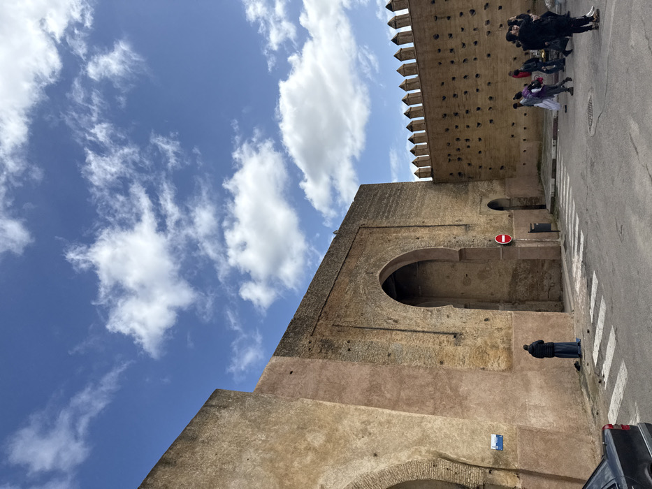 A daytime street scene in Mechouar Fes Jdid, Morocco, depicts a historic city wall and gateway under a partially cloudy blue sky. The ancient reddish-brown wall, constructed of varying stone, features multiple large arched entrances. On the left, a person wearing a dark djellaba and a black puffer jacket is performing prayer on a blue mat, facing an arched alcove in the wall. A black sedan is partially visible in the bottom-left corner. To the right of the praying individual, a red circular "No Entry" sign is mounted on a pole, with a black trash receptacle beneath it. Further to the right, the wall extends with a crenelated top and a section punctuated by numerous small circular indentations. A group of approximately ten individuals, dressed in a mix of traditional and modern attire, gathers on the right side of the paved street near a vendor's display of produce. One individual in black clothing is observed photographing the scene with a smartphone. White crosswalk stripes are visible on the asphalt road in the foreground.