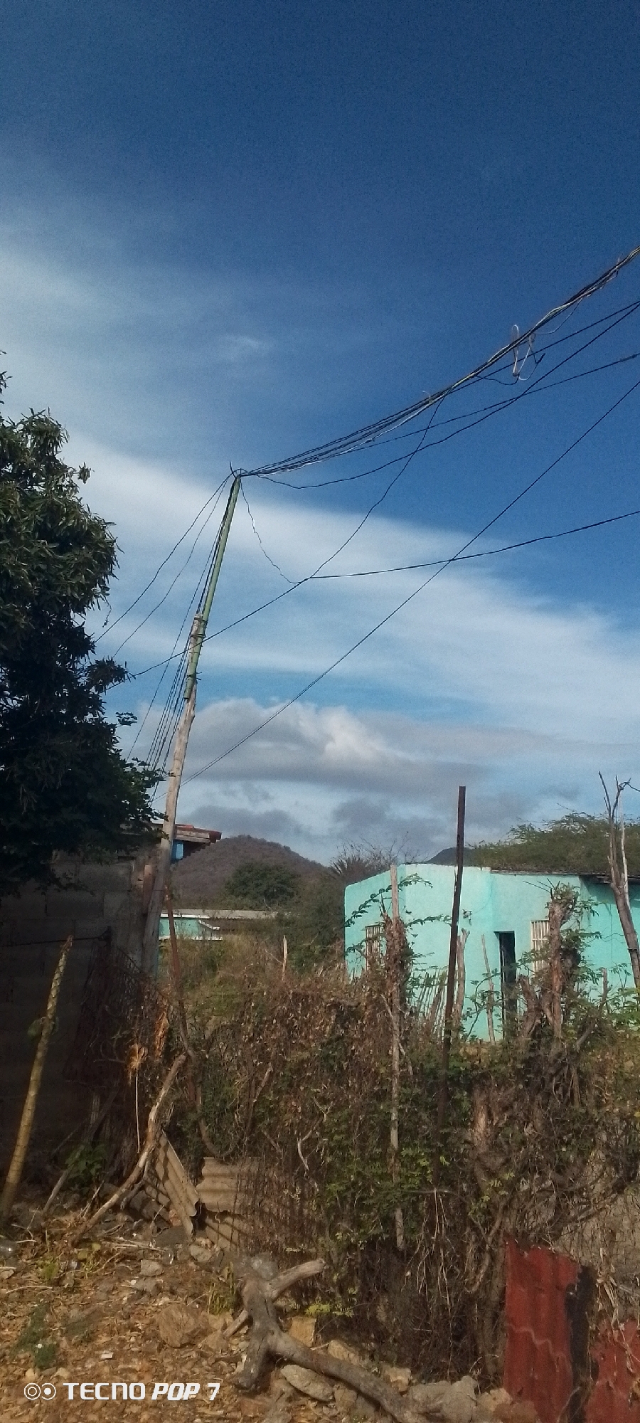 The image depicts a rural scene in Altagracia, Venezuela. The sky is blue with scattered clouds. Power lines are strung across the upper portion of the frame, supported by a makeshift wooden pole. A light-blue, single-story building with a visible doorway and window is partially obscured by dense, dry vegetation. A small, dry, shrub-covered mountain is in the background. In the foreground, there is a mix of dried brush, debris, and a few stones. A partial brick wall is seen on the right, and a concrete block fence is on the left. A tree with green leaves is also on the left. At the bottom of the image, the "TECNO POP 7" watermark is visible.