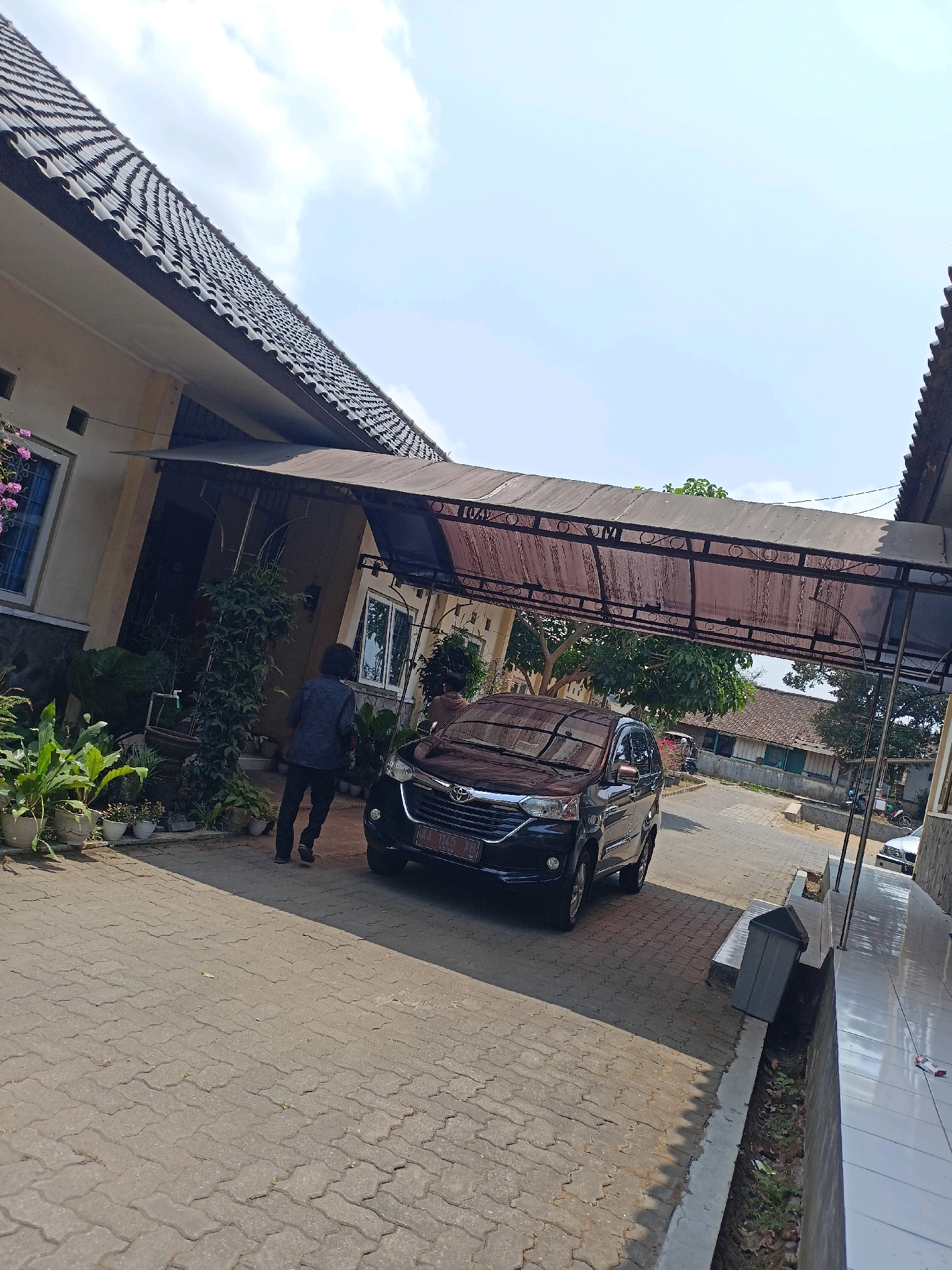 A low-angle, full outdoor shot captures a residential area on a sunny day. In the foreground, a person walks away from the camera towards a dark sedan parked on a paved driveway. The car, a MPV, is positioned under a covered walkway supported by metal poles, its license plate reading "AA 1125 XB".
The driveway is paved with interlocking gray concrete blocks, with a drainage ditch running along its right side, containing some debris and greenery. To the left of the driveway, a cluster of potted plants and lush green foliage are visible. Behind the plants, the side of a light-colored house is visible, with windows and a dark tiled roof. The covered walkway extends from the house and is constructed from a translucent, brownish material supported by a dark metal frame.
In the background, a narrow street is visible, lined with more houses and trees. The sky above is a clear, pale blue with scattered white clouds. The overall scene depicts a typical residential setting in Kewaluhan, Indonesia, characterized by its warm climate and architectural style.