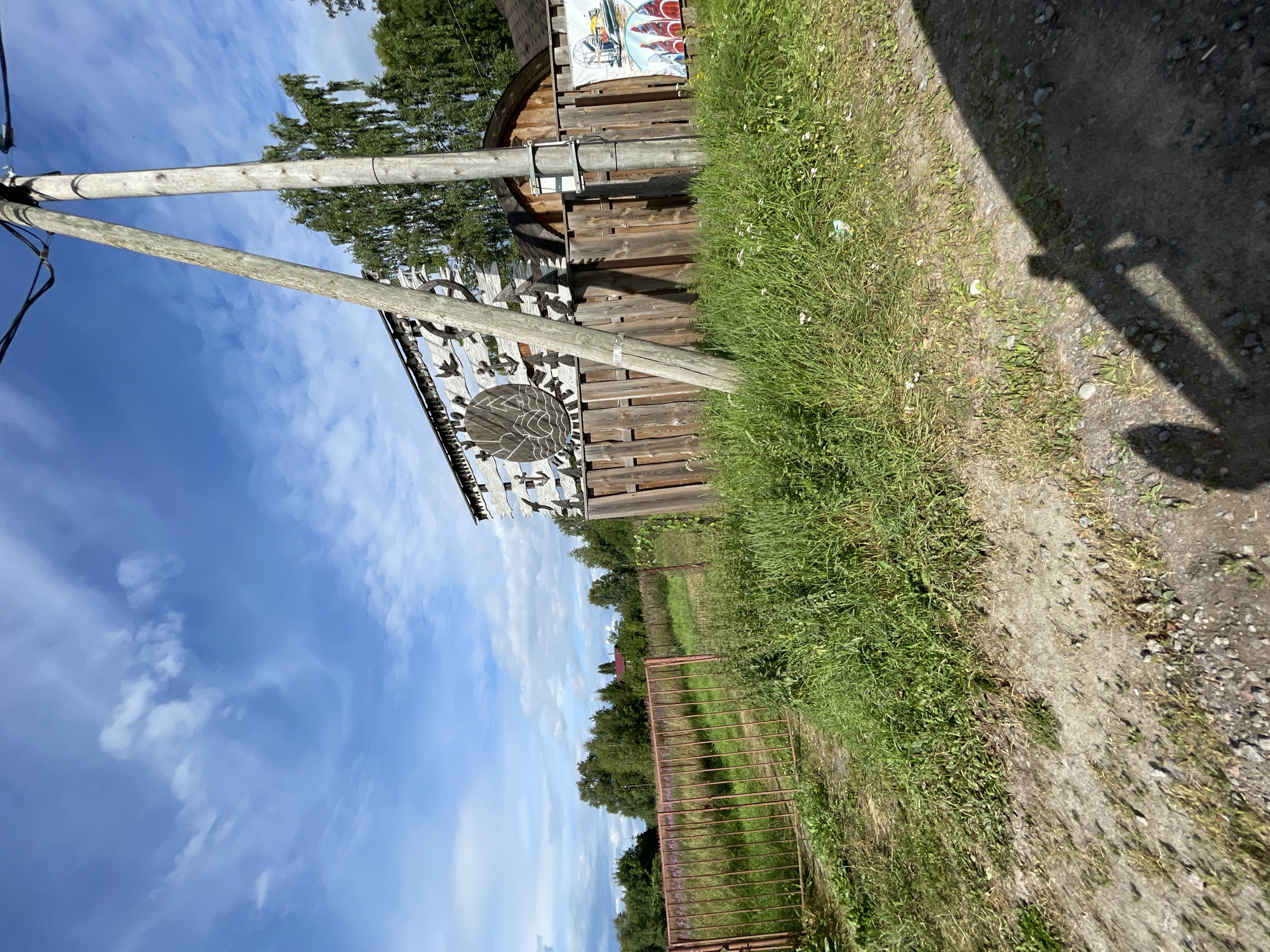 The image depicts a rural scene in the city of Berozovo, Russia, on a sunny day with scattered clouds. In the foreground, a dirt path leads into a grassy area with a rusted red metal gate. Beyond the gate, a wooden fence stretches across the image, adorned with a circular wooden carving featuring waves and an anchor motif, as well as smaller carved nautical symbols like anchors and birds. Behind the fence, a wooden building with a rounded roof is visible. To the right of the fence, a poster or mural is attached, showcasing an airboat and several colorful kayaks or canoes. A tall wooden utility pole with wires runs vertically through the scene, casting a shadow on the ground. The surrounding environment is characterized by lush green grass, trees, and a field with purple flowers in the distance. The overall impression is one of a rustic, possibly recreational, location in a natural setting. There are no people directly visible in the image, only the shadow of a person holding a phone or camera in the lower right corner.