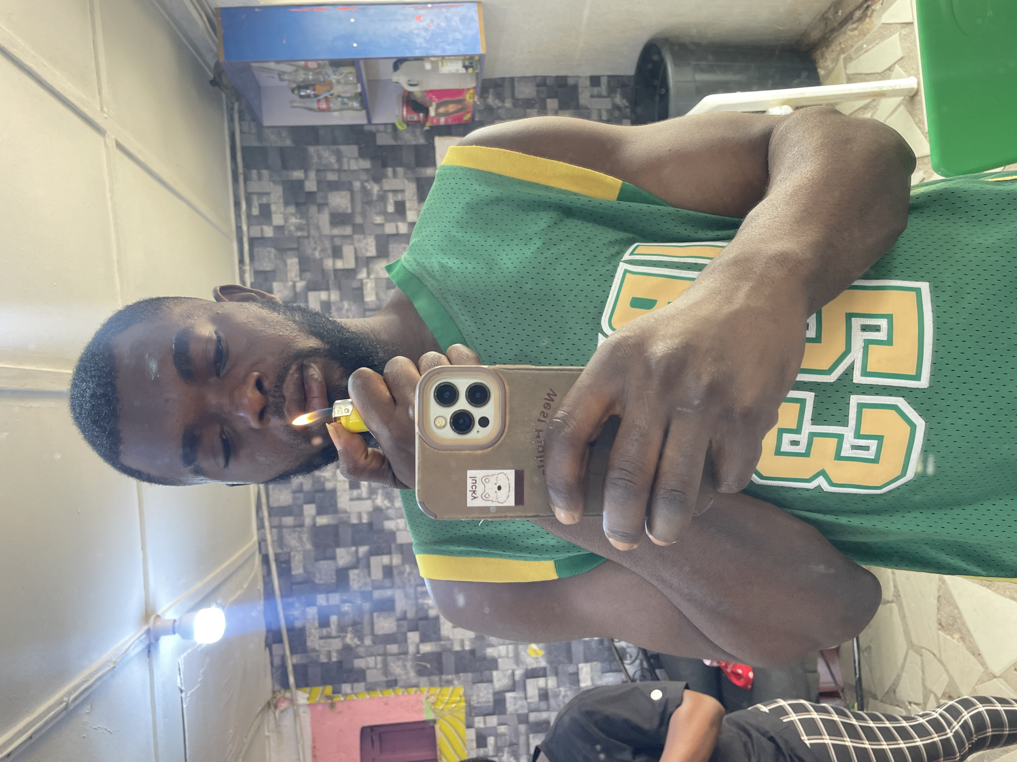 A young Black man with a beard is captured in a mirror selfie indoors. He is holding a yellow lighter with an active flame near his mouth, and his eyes appear closed or squinting. He is wearing a green basketball-style jersey with yellow trim, featuring a prominent yellow and white number "33" on the front.
He holds a light brown or beige smartphone, identified as an iPhone by its triple camera module, in his right hand. The phone case has a white sticker depicting a small bear-like character and the partial word "luck". Mirrored text is also visible on the phone case, but it is difficult to decipher fully.
The background, visible through the mirror's reflection, consists of a tiled wall with a geometric pattern of dark and light squares. Above this, there is a lighter, plain wall with a visible light fixture and a blue shelf holding various bottles, including one clearly identifiable as Coca-Cola. In the lower reflection, a person's legs wearing black and white checkered pants are partially visible. The scene suggests an indoor setting, possibly a commercial establishment like a shop or barbershop, located in Jos, Nigeria.