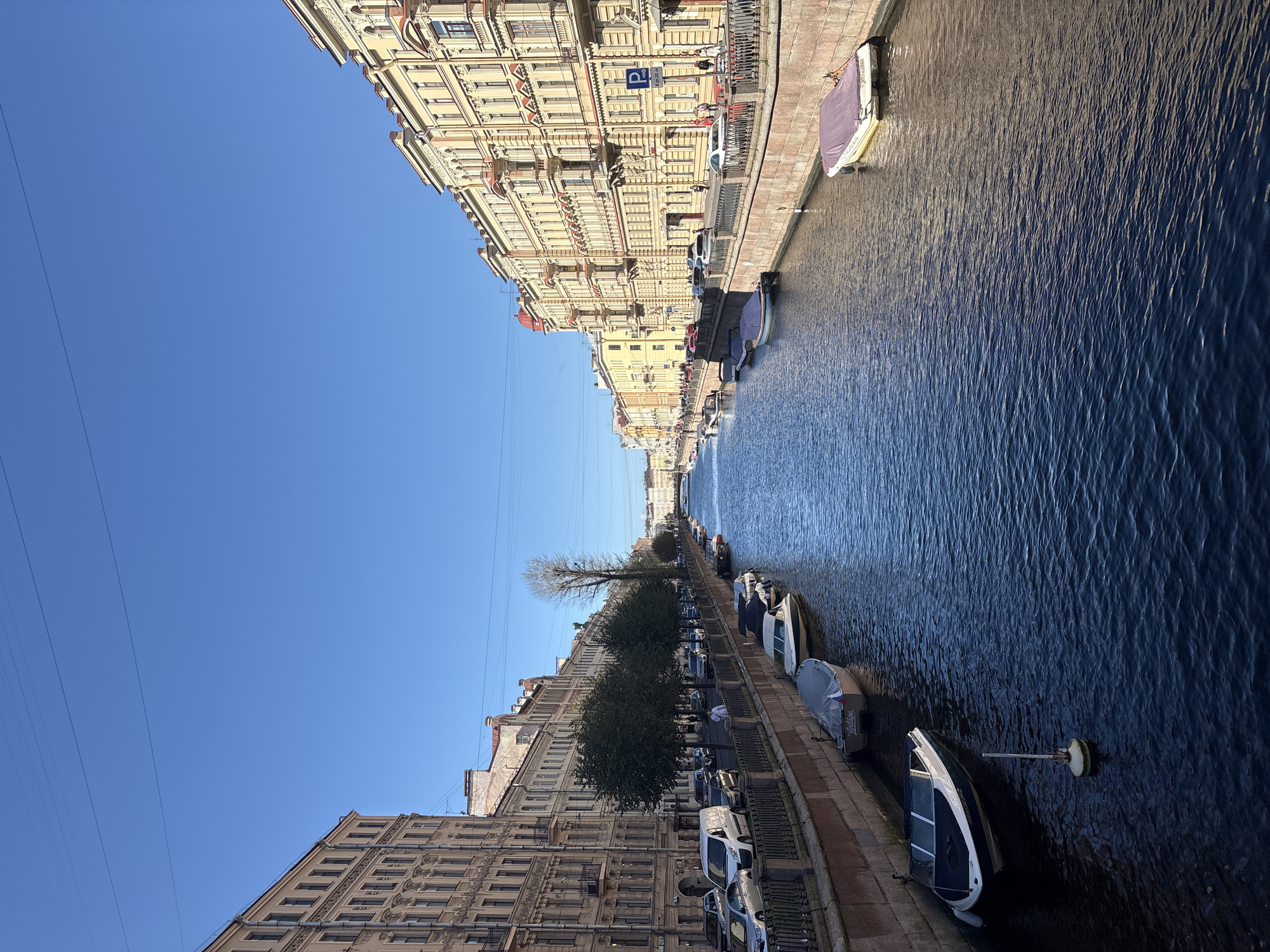 The image captures a scenic view of a canal in Saint Petersburg, Russia, likely the Griboyedov Canal or a similar waterway. The canal is lined with historic, multi-story buildings with ornate facades and large windows, characteristic of the city's architecture. The buildings on the left are taller and appear to be residential or office spaces, while the buildings on the right are a pale yellow and also exhibit classical architectural elements.
Along the canal's banks, there are stone retaining walls and metal railings. Several boats are moored along the edges of the canal, some covered with tarpaulins, suggesting it is a peaceful, possibly late afternoon or early morning scene. On the right bank, a sidewalk is visible with a few pedestrians and parked cars. A sign with a "P" symbol and some Russian text indicates parking is available. Trees are planted along the sidewalks, adding greenery to the urban landscape.
The water in the canal is a deep blue, reflecting the clear, bright blue sky above. The sky is largely unobstructed, with only a few thin power lines visible across it. The overall impression is one of a well-maintained, picturesque European city waterway.