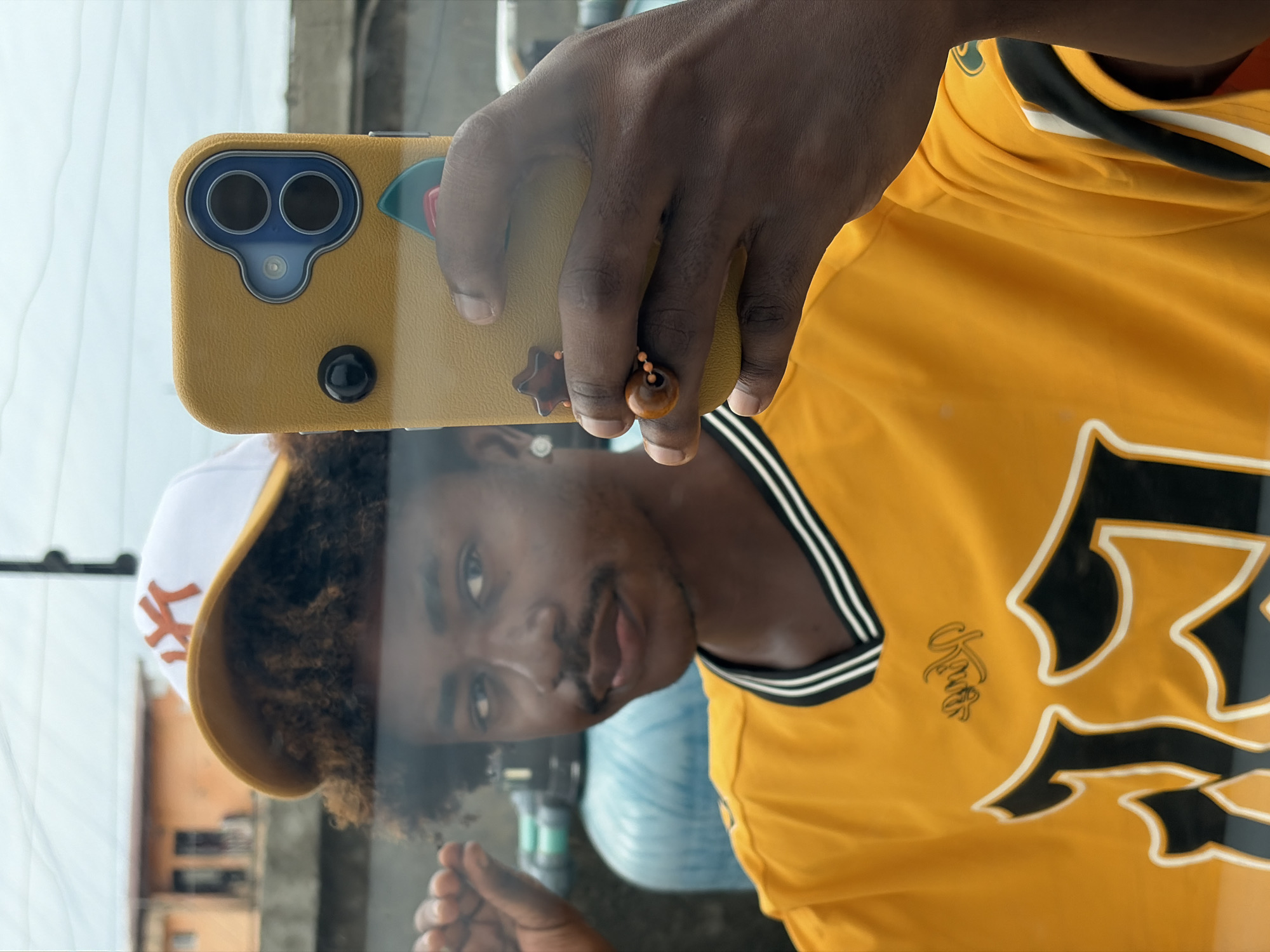 The image shows a person taking a selfie in Igboefon, Nigeria. The person is wearing a yellow jersey with black and white accents, and a white New York Yankees baseball cap with an orange logo. The person is holding a phone with a yellow case decorated with accessories. The reflection in the glass shows buildings and an overcast sky. In the reflection, a partially visible blue container, possibly a water jug, sits near the person. The person's facial expression is neutral.