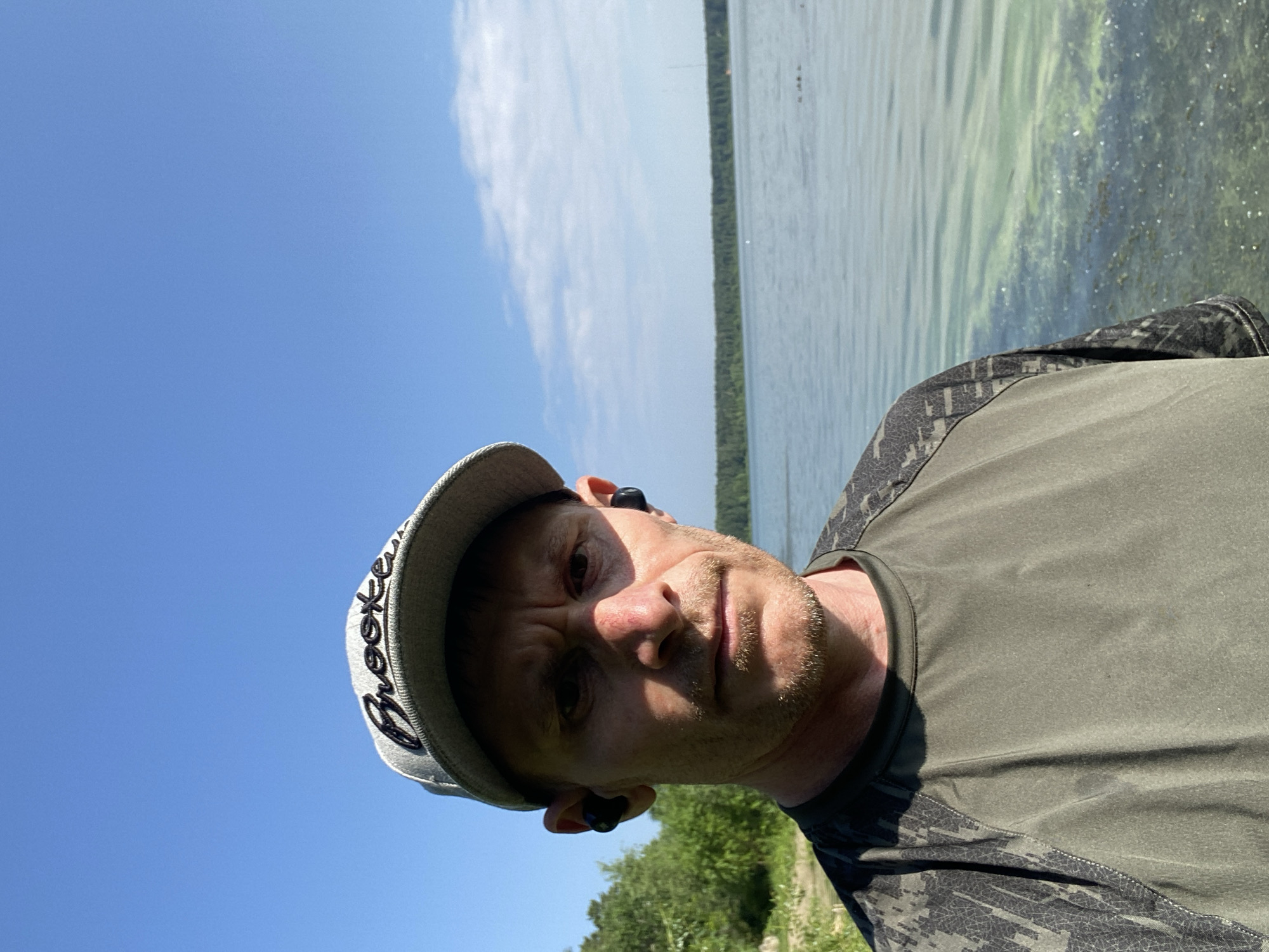 A man wearing a gray baseball cap with "Brooke" embroidered on it and black wireless earbuds stands in front of a lake. He is wearing a dark green t-shirt with a camouflage pattern on the sleeves. The sky is clear and blue with a few scattered clouds. The lake is calm and reflects the sky. Trees line the far bank of the lake. The image appears to have been taken outdoors on a sunny day. There is no indication that this image is located in the city of Yekaterinburg, Russia.