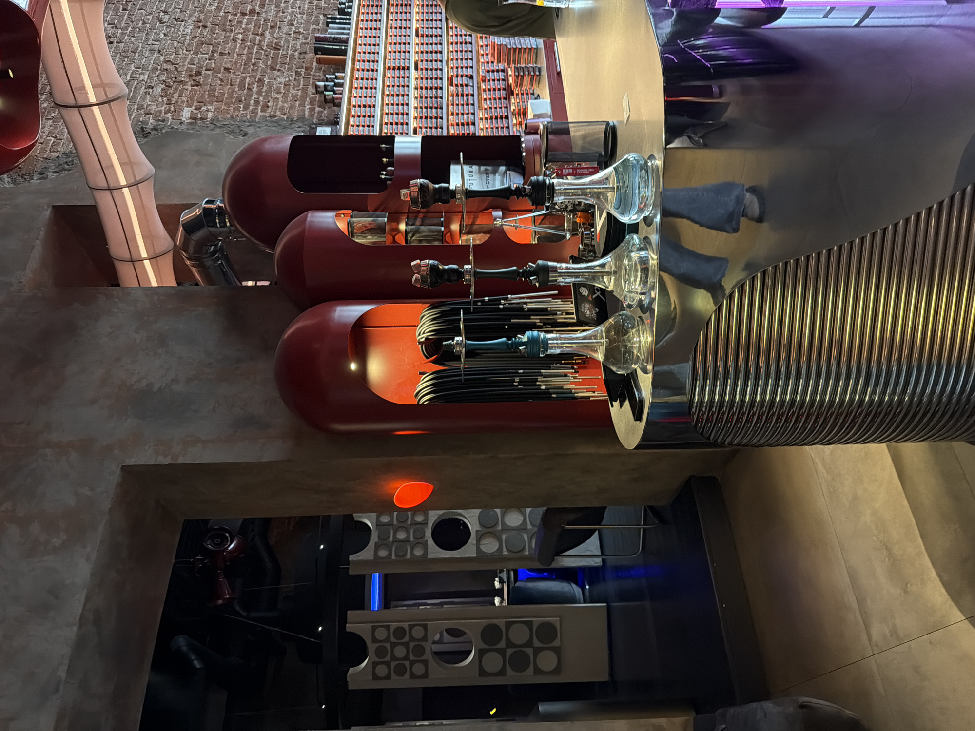 The image shows the interior of the Butler restaurant in Moscow, Russia. The scene includes a view of the restaurant's decor, featuring red capsule-shaped elements containing hookah apparatus. The capsules are mounted on a metallic structure. A stylized pipe resembling bamboo is also visible, alongside a wall made of red brick. Below the hookah installations, there is a glimpse into a darker, interior space, with metal shelves.
