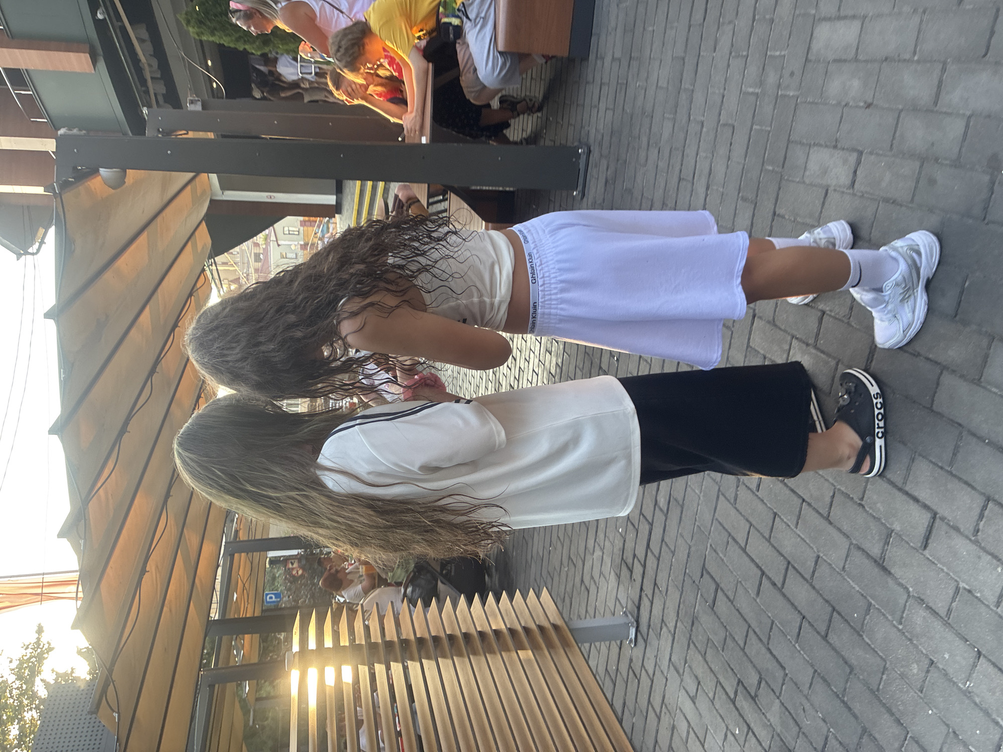 The image captures an outdoor scene, possibly at a cafe or restaurant patio, with two young females standing back-to-back. The female on the left, viewed from behind, has long, light brown hair and wears a white t-shirt with black stripes on the sleeves and black culottes. She is wearing dark Crocs. The female on the right, also viewed from behind, has long, dark, curly hair and is wearing a white tank top and a white skirt. She is wearing white sneakers and white socks. The background shows outdoor seating with wooden slatted dividers and a large awning providing shade. Other people are visible in the background sitting at tables. The setting appears to be urban, with some buildings visible in the distance. The sun is low in the sky, casting long shadows and creating a warm, golden light. The ground is paved with rectangular bricks. No specific landmarks indicate the location as Sochi, Russia.