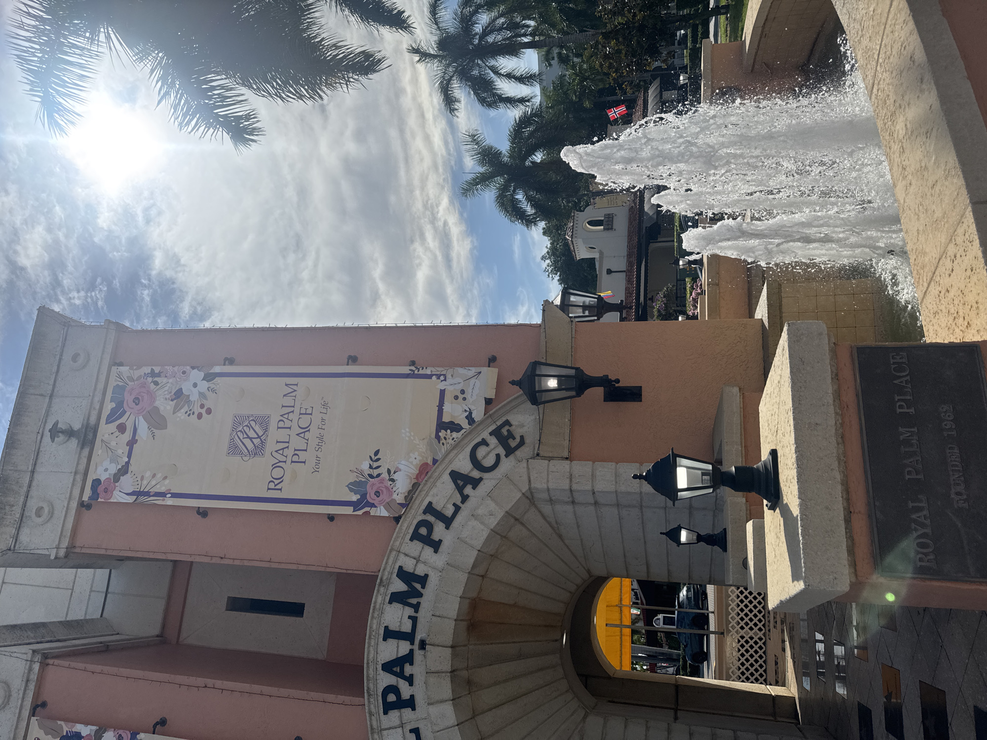 The image depicts the entrance of "Royal Palm Place" in Boca Raton, Florida, United States of America. The architectural style is Mediterranean Revival, characterized by its stucco exterior, arched entryway, and decorative banners. The sun shines brightly, casting a warm glow on the scene, with palm trees visible in the background. A multi-tiered fountain stands in the foreground, its water jets creating a dynamic visual element. The name "Royal Palm Place" is prominently displayed on the arched entrance and on a bronze plaque at the base of the fountain, which also indicates the founding year of 1962. The overall impression is one of a well-maintained, upscale commercial or retail space.