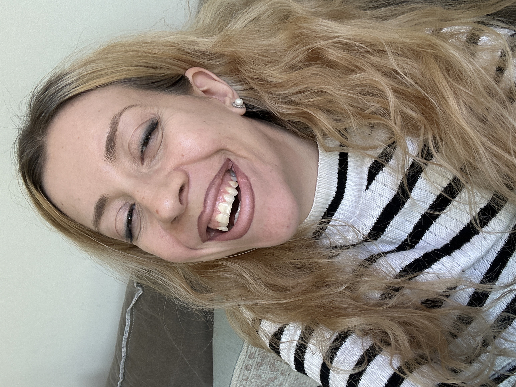 A smiling woman with long, wavy blonde hair is captured in a close-up, head-and-shoulders portrait, tilted at an angle. She is looking towards the viewer with a wide, joyful smile that crinkles the skin around her eyes and reveals her upper and lower teeth. Her right eye is slightly more visible, showing dark eyeliner and mascara. She has light-colored eyebrows.
She is wearing a white sweater with horizontal black stripes. A pearl earring set in a silver-toned surround is visible in her right ear. Her blonde hair is layered and appears to have some darker roots.
The background consists of a light, neutral-colored wall on the upper left side, transitioning to a soft, textured fabric, possibly a cushion or blanket, in shades of grey and beige, on the lower right. The overall impression is an indoor, relaxed setting, likely a casual photoshoot or selfie. There is no visible text in the image. The woman conveys an emotion of happiness and cheerfulness.