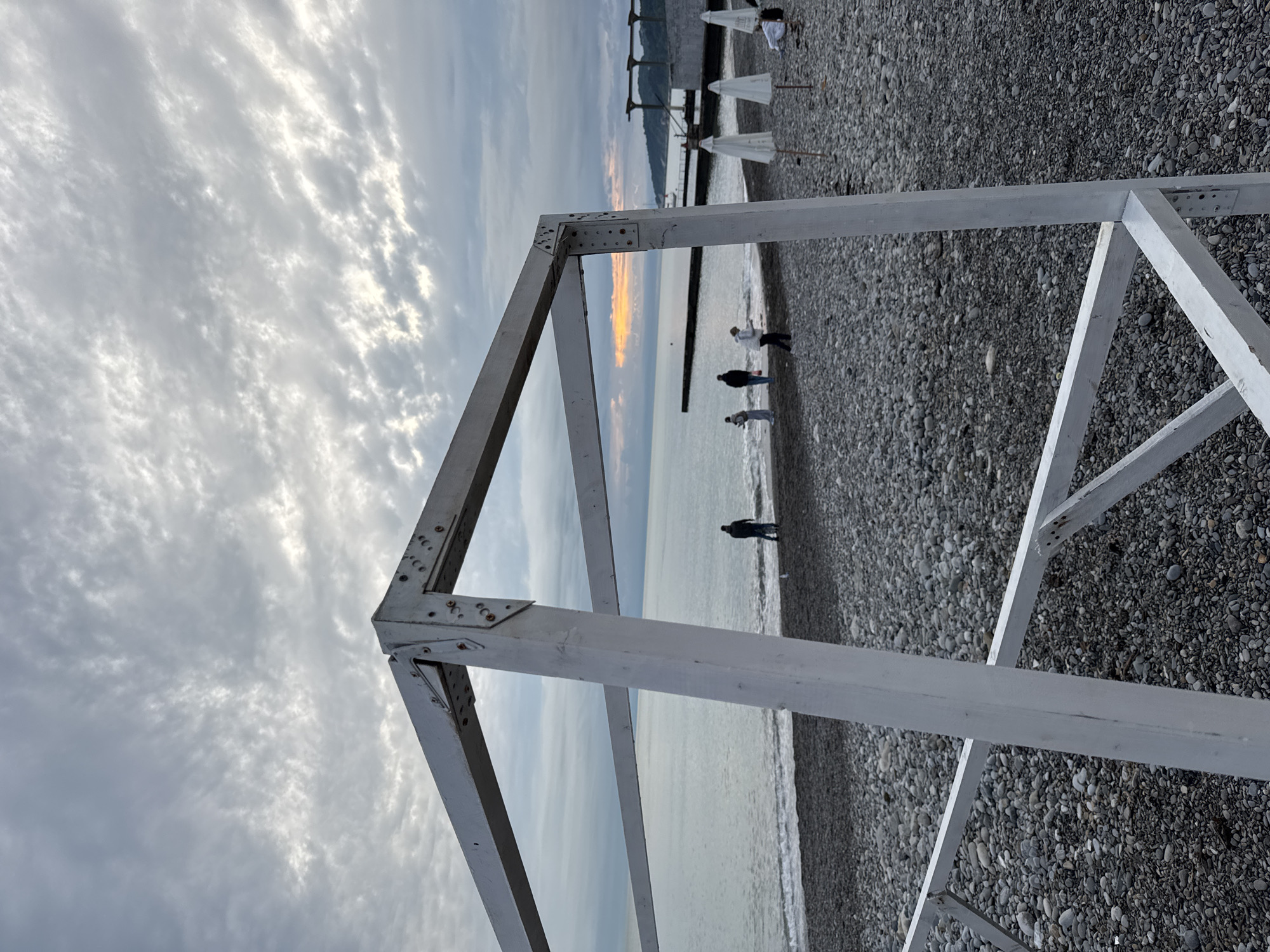 The image captures a tranquil coastal scene in Sochi, Russia, likely during sunrise or sunset, characterized by soft, diffused light. The perspective is low, looking through a large, white, geometric frame that dominates the foreground. This frame, appearing to be constructed from painted wood or metal, features bolted connections at its joints and adds a structural element to the composition.
Beyond the frame, a wide pebble beach extends towards the sea. Several individuals are visible in the mid-ground, scattered along the water's edge, seemingly strolling or standing. The sea is calm, reflecting the sky's gentle light. A notable detail is the warm, orange and yellow glow on the horizon over the water, indicating the sun's position and contributing to the serene ambiance.
The sky is largely covered with light, wispy clouds, allowing some brighter patches of light to filter through. In the distance, behind the beach, mountains or hills are subtly outlined against the horizon. Further down the beach, several white, folded beach umbrellas are visible. The overall mood is peaceful and contemplative. There is no visible text in the image.