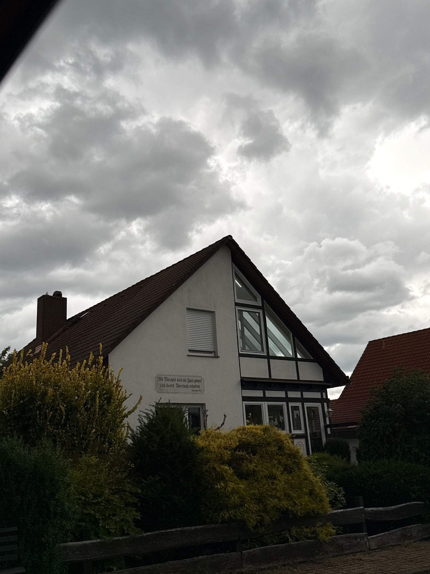 A white house with a dark tiled roof and black timber framing is pictured in a cloudy, overcast environment. A brick chimney protrudes from the left side of the roof. The house has several windows, including a large, triangular window on the upper left side. A sign with text is affixed to the exterior wall. The house is partially obscured by lush green and yellow bushes in the foreground. A wooden fence runs along the bottom of the image. The image is located in Bad Sachsa, Germany.