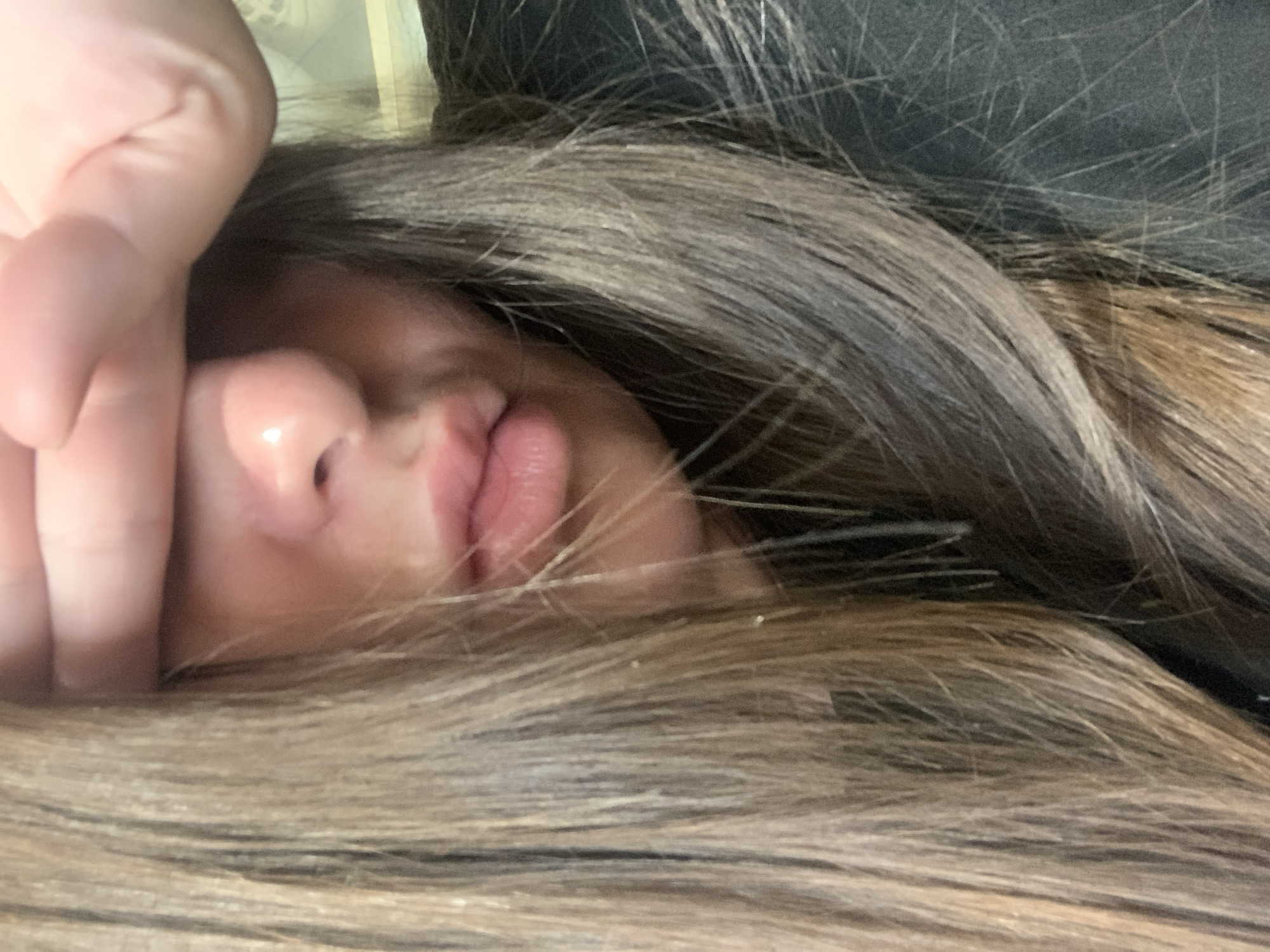 A close-up, indoor shot shows the lower half of a person's face, with their hand covering their eyes. The person has long, brown hair that falls around their face and shoulders. Their lips are slightly parted, and they appear to be wearing a black garment. The lighting is soft, and the background is blurred, suggesting an out-of-focus environment.