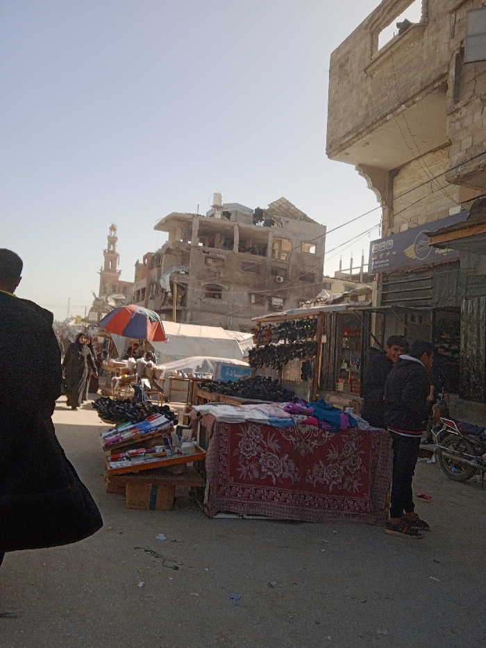 The image captures a bustling street scene in Khan Yunis, Palestinian Territory, under clear daylight. In the immediate foreground on the left, the back of a person in dark clothing is partially visible, facing inward towards the scene. The ground is an unpaved or dirt-covered thoroughfare with scattered refuse.
In the mid-ground, market activity is evident. On the right, a display table covered by a red rug with a floral pattern presents folded textiles. Adjacent to it, two young males stand, one in a dark jacket facing the viewer, beside a parked motorcycle. To their left, a stall displays numerous dark shoes suspended from a frame. Another stall is partially shaded by a red, blue, and white striped umbrella, showing merchandise including stacks of books or magazines on wooden stands. Additional market stalls with white tarpaulin coverings extend into the mid-distance. Multiple individuals, including a woman in a dark abaya, are observed walking amongst the stalls.
The background is dominated by architectural structures. On the left, a light-brown, ornate minaret of a mosque rises prominently. Central to the frame, a multi-story concrete building shows significant structural damage, with exposed rebar, missing wall sections, and visible utility elements like water tanks and solar panels on its roof. To the right, a partially constructed or damaged building façade features exposed concrete blocks, an ornamental arched element, and a blue shop sign with white Arabic text and an emblem. The sky is light blue and clear.