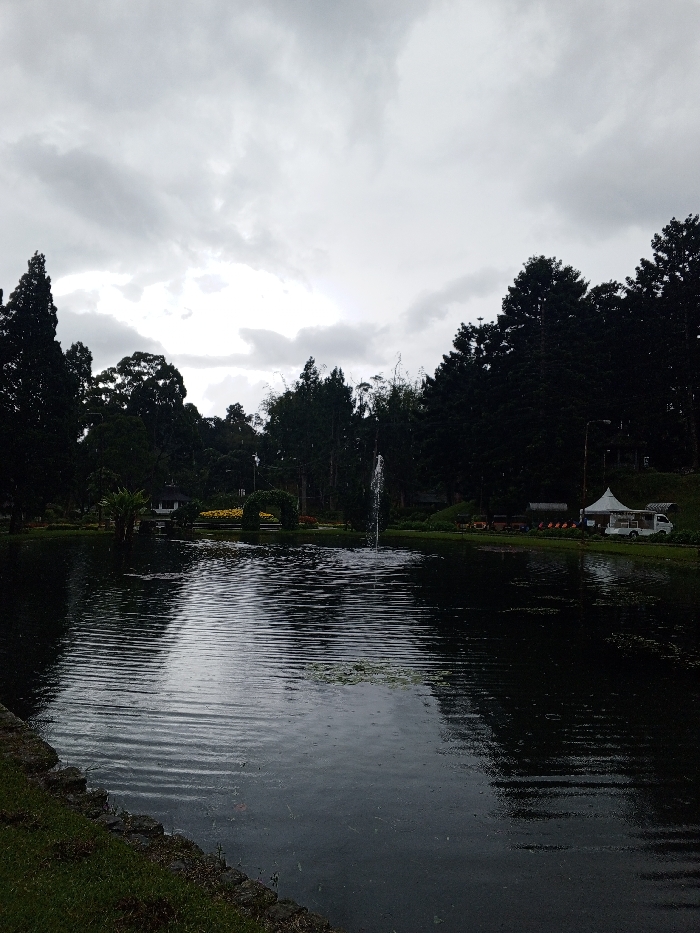 A serene, cloudy day at a park. A dark, reflective lake with a central fountain is surrounded by lush, dark green trees. An arched floral decoration sits on a small island within the lake. A white tent-like structure and van are visible on the far bank. Raindrops pockmark the water's surface, indicating a light shower. The foreground shows a rocky edge with grass.