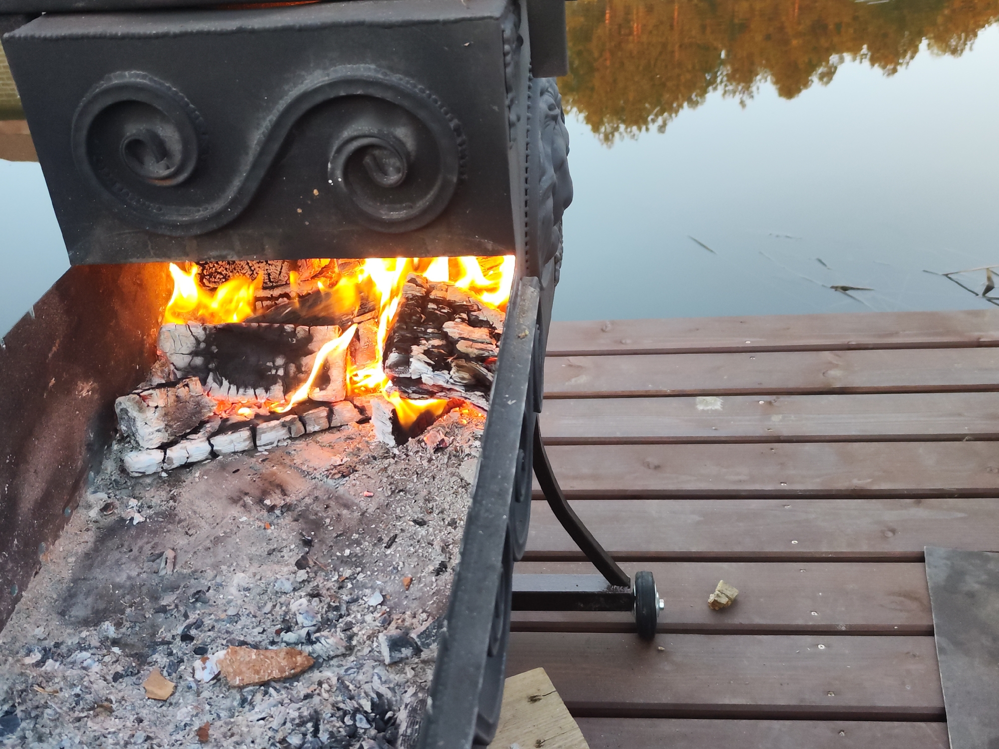 A metal brazier with a fire burning inside sits on a wooden deck next to a body of water. The brazier is ornate, featuring spiral designs and what appear to be sculpted lion heads. The fire consists of burning logs and glowing embers. The deck is made of wooden planks, and a small wooden block is visible under the brazier's wheel. Beyond the deck, water stretches out, reflecting the trees on the opposite shore. The location is Chaikovsky, Russia.