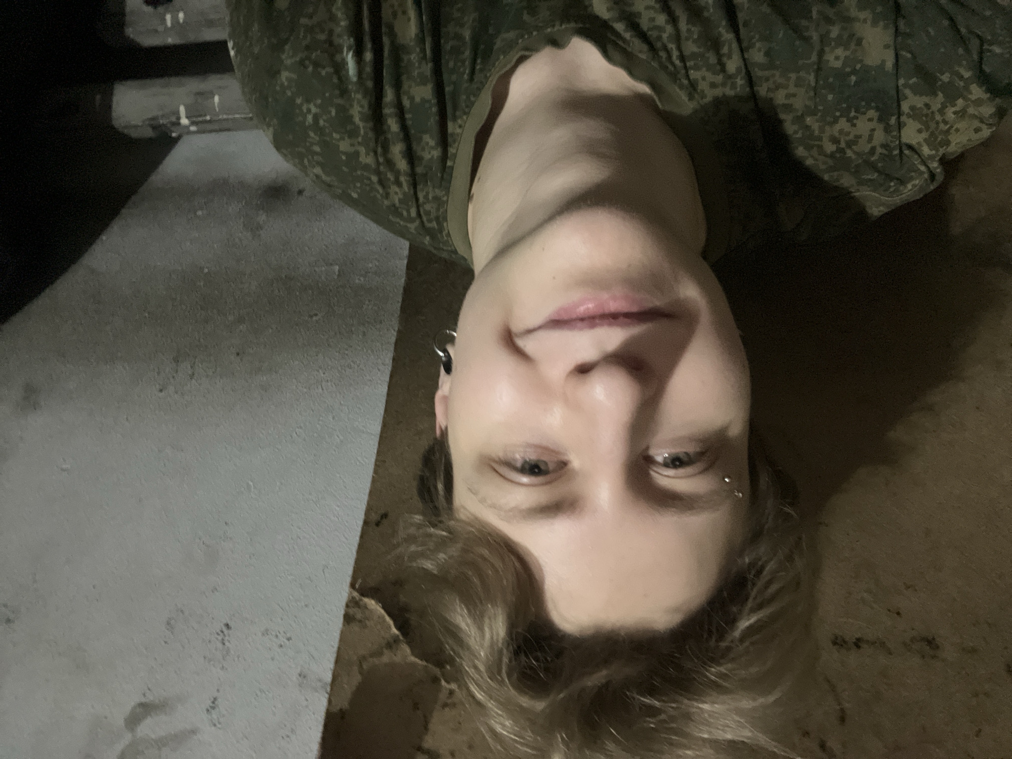 A selfie of a person with blond hair wearing a camouflage shirt. The person is located indoors with a concrete wall in the background. The location is identified as the city of Chaikovsky, Russia.