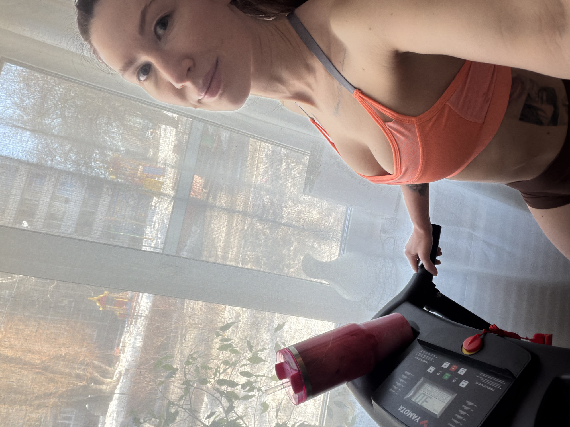 The image depicts a woman inside a residence, standing beside a black "YAMOTA" brand treadmill, taking a selfie. She wears an orange sports bra with grey straps and dark brown shorts, displaying tattoos on her upper chest and left hip. Her right hand rests on the treadmill's handle. A red tumbler with a straw containing a red liquid is positioned in the treadmill's cup holder. A white, textured, pear-shaped decorative object is on the windowsill to the right of the tumbler. Through the sheer white window curtains, an urban residential courtyard is visible. The ground outside is covered in snow, and bare deciduous trees are present. In the mid-ground, a distinctive red and yellow pagoda-style playground structure is visible. Multi-story apartment buildings constitute the background. The outdoor environment and the specific playground structure indicate the scene is located in a residential courtyard, consistent with the area around Ulitsa Krasnopiterskaya, 24A, Perm, Perm Krai, Russia. Bright sunlight enters the room from the window.