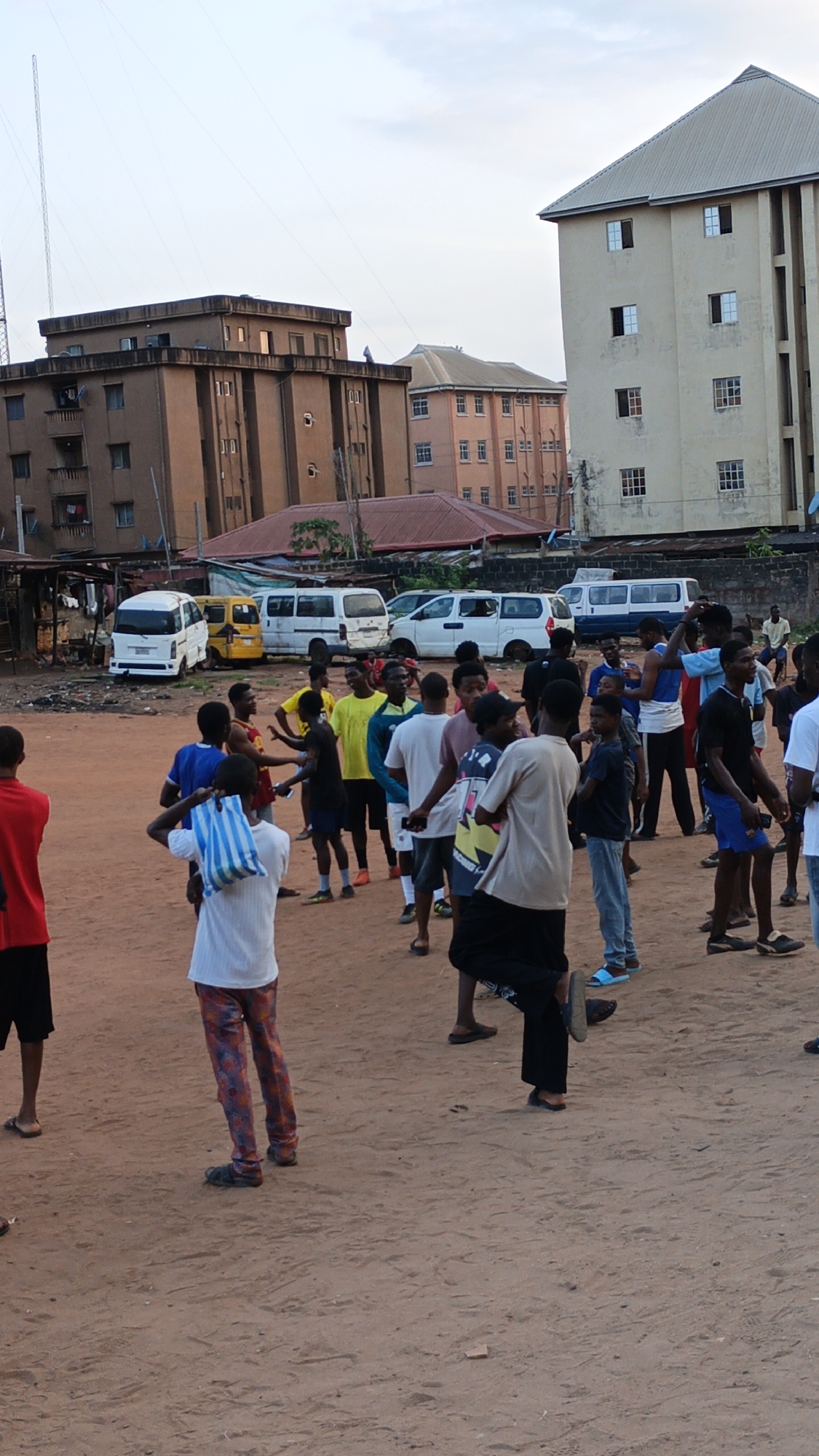 The image captures a vibrant gathering of young men and boys on a reddish-brown dirt field, likely in Onitsha, Nigeria. The group, consisting of approximately twenty individuals, is scattered across the open space, some appearing to be engaged in conversation, while others stand casually, suggesting an informal social event or preparation for a recreational activity, possibly football, given some are wearing athletic attire.
In the background, an urban landscape unfolds with several multi-story buildings. To the left, a prominent brown building with multiple windows and balconies stands, next to which a tall, slender communication antenna reaches towards the sky. On the right, a lighter-colored building with a pitched roof and visible window frames dominates the skyline. Other structures and rooftops are also visible between these main buildings. Behind the gathering, a row of parked white and yellow mini-vans or buses indicates a nearby road or parking area.
The overall lighting suggests late afternoon or early evening, with an overcast sky casting a soft, diffused light over the scene. The ground is bare earth, marked with footprints and minor irregularities. One young man in the foreground, facing away, wears a white t-shirt and colorful patterned trousers, carrying a blue and white striped bag over his shoulder. Another figure is partially visible on the far left, dressed in a red t-shirt and dark shorts.
The scene conveys a sense of community and everyday life in an urban Nigerian setting, characterized by the informal gathering of youth against a backdrop of local architecture and infrastructure. No explicit text or signage is clearly visible in the image.