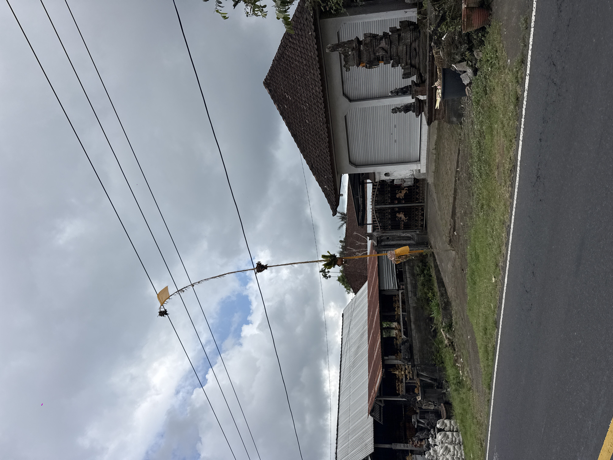 The image captures a roadside scene in the city of Susut, Indonesia, under a cloudy sky. A tall, slender pole with a yellow flag and some decorative elements stands prominently, reaching towards power lines that stretch across the frame. To the left, a long building with a corrugated metal roof, possibly a workshop or storefront, displays stacks of white bags and various items, some appearing to be sculptures or crafts, on shelves. In the center, a gate with ornate metalwork leads to a building with a tiled roof and two large, gray roller doors. To the right of this building, a stone shrine or sculpture stands in front of a gray wall, adorned with smaller stone figures. A dark bin and debris are present in front of the shrine. The foreground shows a paved road with a white line marking its edge and a yellow line on the far left. The environment appears rural or semi-urban, with vegetation and palm trees visible in the background.