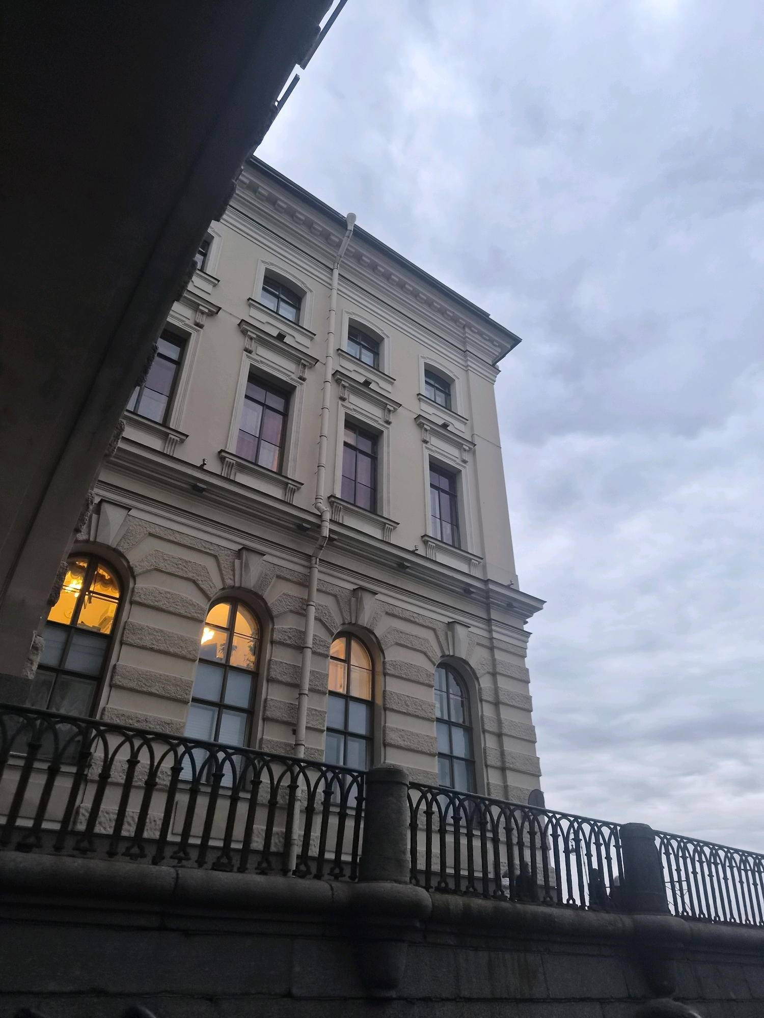 The image captures a portion of a pale yellow, multi-story building in Saint Petersburg, Russia, observed from a low angle. The building features classical architectural elements, including a decorative cornice, pilasters, and recessed window frames. Several windows are visible, some displaying a warm, orange-yellow light from within, suggesting interior lighting. The sky above is overcast with a grey, cloudy texture. In the foreground, a dark, ornate metal railing with arched elements runs horizontally across the frame, positioned above a stone embankment. A dark, solid structure, possibly a bridge or overpass, partially obscures the upper left portion of the building and the sky. The scene depicts an urban architectural setting under a cloudy sky, likely in the evening or during a period of low light.