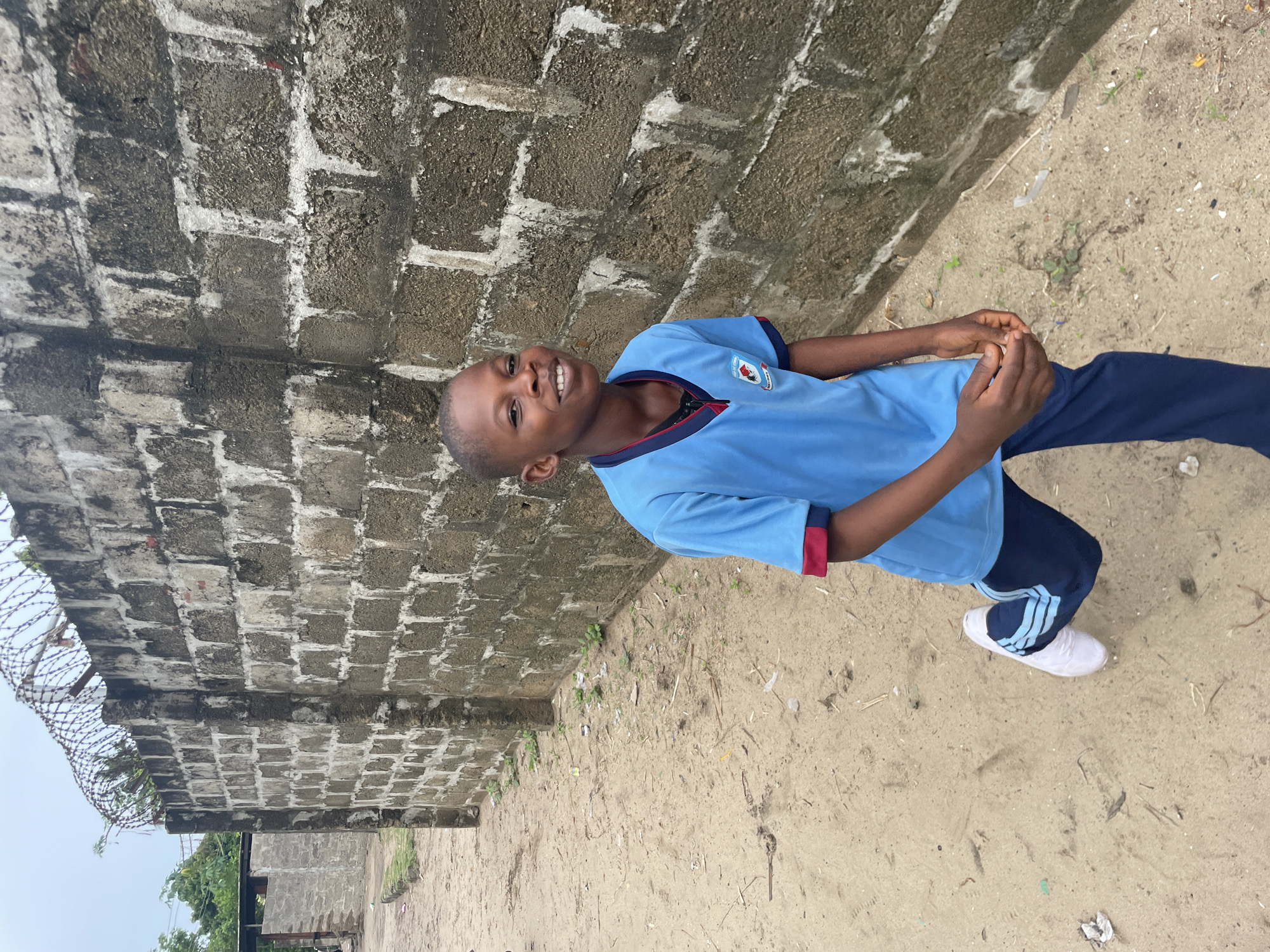 A boy in a light blue shirt and dark blue pants stands in front of a weathered brick wall. The wall has barbed wire along the top edge in the background. The boy smiles, his hands clasped together. The ground is sandy and has a few weeds. A white shoe is visible on his right foot. A building is visible in the background. The location is Badagry, Nigeria.