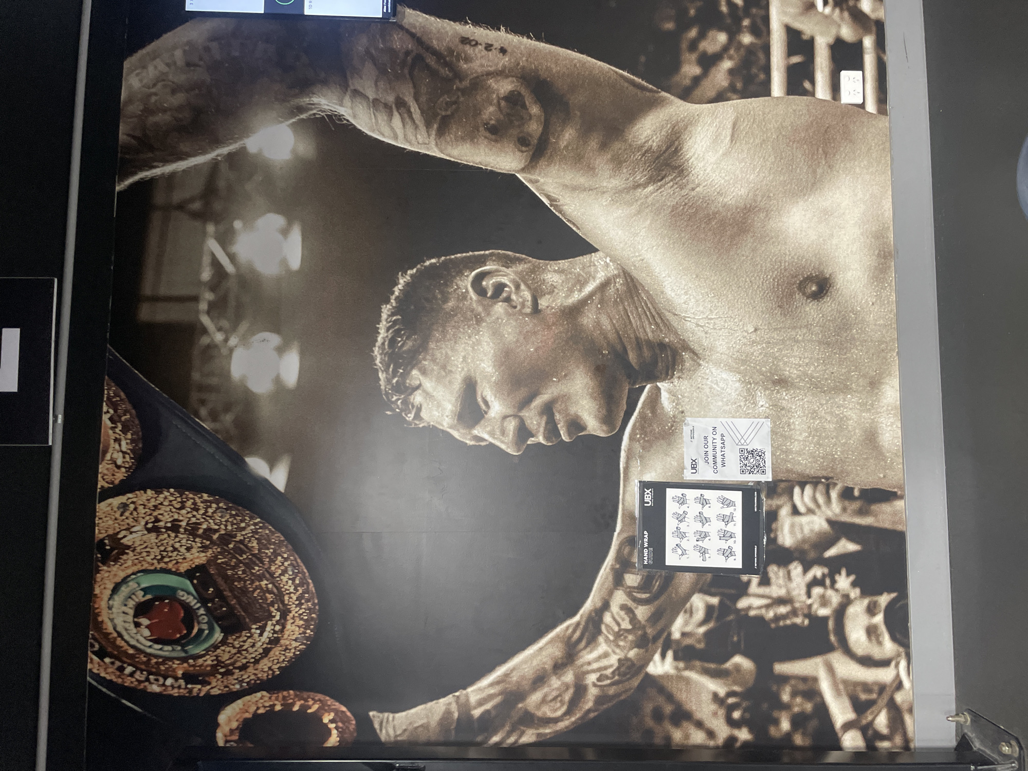 A large mural of a boxer with his arms raised in victory is displayed on a wall. The boxer is muscular and sweating, with tattoos on his arms. He is wearing a championship belt. Two smaller posters are affixed to the mural. One poster is a "HAND WRAP GUIDE" with diagrams of how to wrap hands. The other poster is an advertisement for a UBX community on WhatsApp, including a QR code for joining. The overall tone of the image is celebratory and athletic.