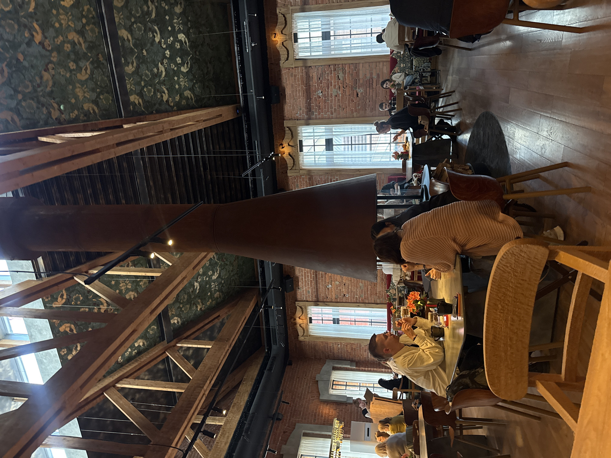 The image depicts the interior of a restaurant with a rustic and industrial aesthetic, likely located in Moscow, Russia. The space features exposed wooden beams supporting a high ceiling adorned with a patterned wallpaper featuring birds and floral motifs. A large, conical chimney or ventilation shaft dominates the center of the room, its reddish-brown surface contrasting with the exposed brick walls and large windows with white curtains.
Several tables are occupied by diners, engaged in conversation and eating. In the foreground, a man in a light-colored shirt is seated at a table, looking towards his companion, a woman wearing a striped cardigan. Other patrons are visible throughout the restaurant, some in the mid-ground and background, seated at various tables. The flooring is made of light-colored wooden planks. The overall ambiance suggests a bustling yet comfortable dining experience.