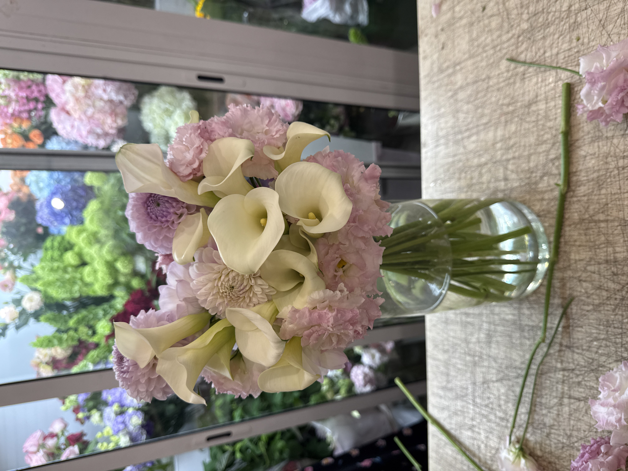 A floral arrangement of white calla lilies, pink carnations, and light purple dahlia flowers in a clear glass vase sits on a textured wooden surface. Behind the vase, through a window, a blurred background displays a vibrant assortment of various colorful flowers. The overall lighting is soft, highlighting the delicate textures of the flowers.