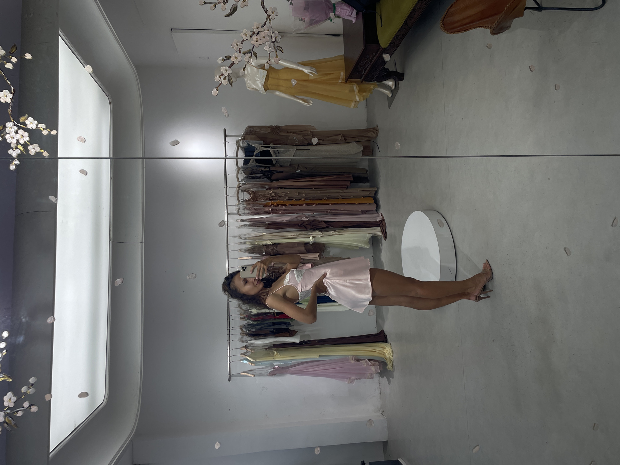 A woman is depicted taking a mirror selfie in a clothing boutique in Thủ Đức, Vietnam. She stands in a full-body side pose, facing her reflection, with her head turned slightly towards the camera and a subtle smile. She has long, dark, wavy hair and is wearing a light pink, short satin slip dress with white lace details on the bust. Her right hand rests on her hip, and her left hand holds a smartphone with a light-colored case up to her face, capturing the image. She wears open-toed, light-colored high-heeled mules.
The setting appears to be a modern retail space with plain white walls and a light grey floor. Behind the woman, a metal clothing rack displays numerous garments in various muted tones, including pink, yellow, brown, and grey, suggesting a range of flowing dresses and attire. To the left of the mirror, a white circular pedestal is partially visible on the floor. Scattered across the floor and visible in the reflection are small, light-colored petal-like decorations.
The large mirror reflects the opposite side of the store. A white female mannequin, dressed in a strapless yellow and white gown, stands on the right side of the reflection. Above and to the right of the mannequin, decorative branches with white and pink cherry blossom-like flowers are affixed to the mirror. Adjacent to the mannequin is a dark brown, ornately carved wooden table, on which a bouquet wrapped in purple paper is placed. A green upholstered bench and a brown leather chair with a black metal frame are partially visible to the right of the table in the reflection. A vertical seam divides the mirror approximately down the center of the image. The ceiling features a large, rectangular light fixture, visible in the reflection at the top.