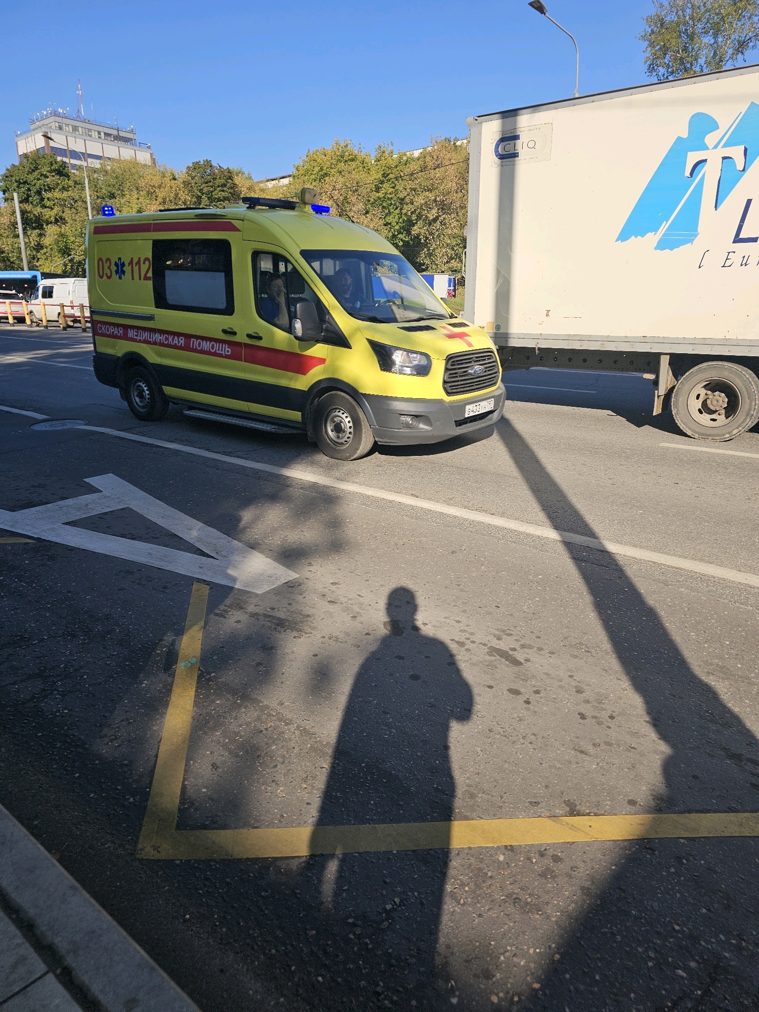 An ambulance is parked on a street in Moscow, Russia, with a large white truck to its right. The ambulance is yellow with red and black stripes and displays the Russian emergency number "03 112" and "СКОРАЯ МЕДИЦИНСКАЯ ПОМОЩЬ" (Emergency Medical Aid). The truck has a logo with blue and white graphics and the text "CLIQ". The street has yellow and white road markings. The sun is casting long shadows of people onto the road. In the background, there are trees and a building with a communications tower on top. The sky is clear and blue.