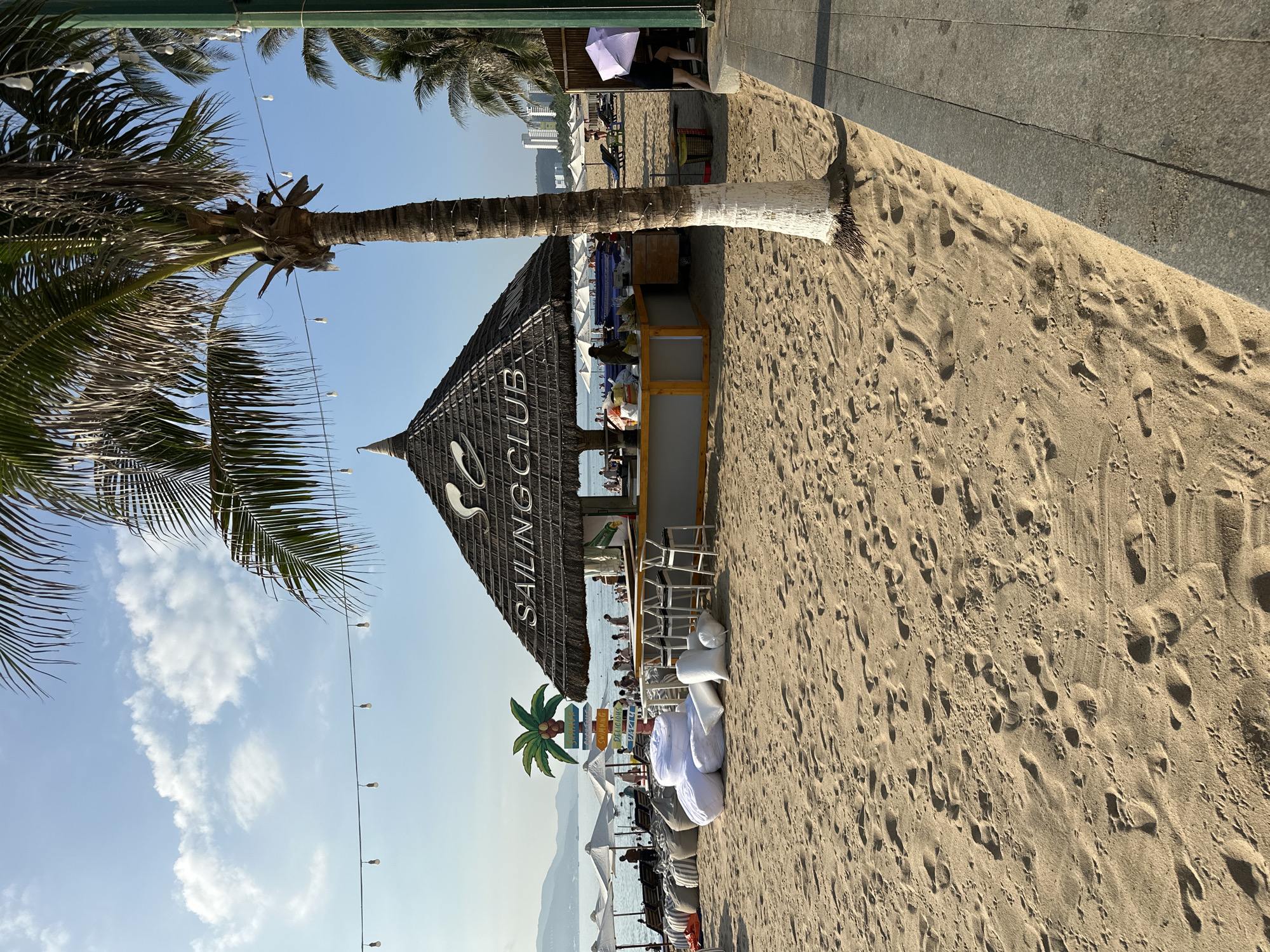 The image captures a beachside scene in Nha Trang Ward, Vietnam. In the foreground, sand is textured with numerous footprints. A palm tree with a white-painted base stands prominently, its fronds extending upwards and towards the left. Behind the palm tree, a thatched-roof beach bar labeled "SAILING CLUB" is visible. The bar has stools and a counter area. Scattered on the sand in front of the bar are several large, white beanbag chairs. Beyond the bar, the beach stretches out with rows of lounge chairs and umbrellas, and a calm body of water is visible in the distance. Several people are present on the beach and in the water. The sky is blue with scattered clouds, and string lights are hung across the scene. To the right of the palm tree, a paved walkway runs alongside the beach.