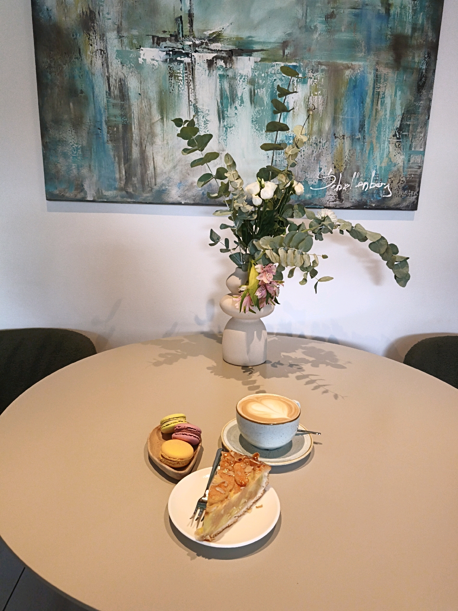 An abstract painting with shades of blue, green, and brown hangs on a white wall above a round, beige table. On the table, a bouquet of eucalyptus and pink flowers is arranged in a white, modern vase. To the right of the vase, a light blue mug with a heart-shaped latte art rests on a saucer with a spoon. In front of the mug, a slice of apple pie sits on a white plate with a fork. To the left of the pie, a small wooden dish holds three colorful macarons: yellow, pink, and orange. The table casts a shadow of the eucalyptus branches on its surface. Two dark green chairs are partially visible on the left and right sides of the frame. The scene is likely from a cafe in Tallinn, Estonia, given the style of the art and decor. No people are visible.
