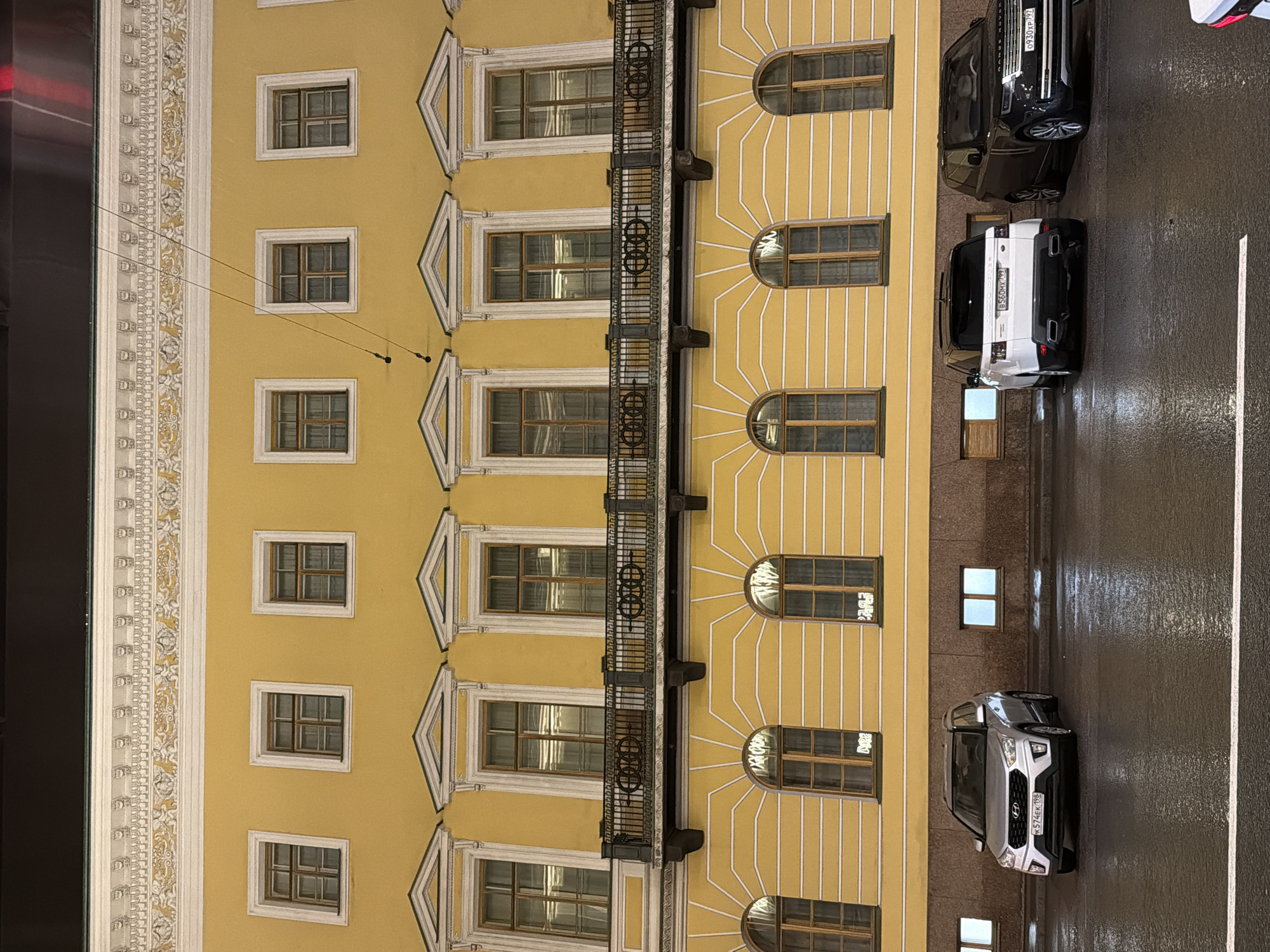 A nighttime, eye-level, wide shot captures a segment of a grand, neoclassical building facade in Saint Petersburg, Russia, characterized by its vibrant yellow paint and intricate white decorative moldings. The building features two distinct rows of windows. The upper row consists of rectangular windows with multiple panes, each framed by white, pediment-like structures. Below this, a decorative balcony with a black metal railing and ornamental circular motifs spans across the facade. The lower row of windows is arched and also divided into panes, with white linear detailing radiating outwards from each window frame across the yellow wall. The street in front of the building is wet and reflective, indicating recent rain, with a white line marking a lane. Three SUVs are parked on the wet asphalt. From left to right, the first is a silver Hyundai Creta, followed by a white Range Rover Sport, and finally a black Range Rover. Their headlights and the building's illuminated windows cast reflections on the wet ground. The sky above is dark, suggesting late evening or night.