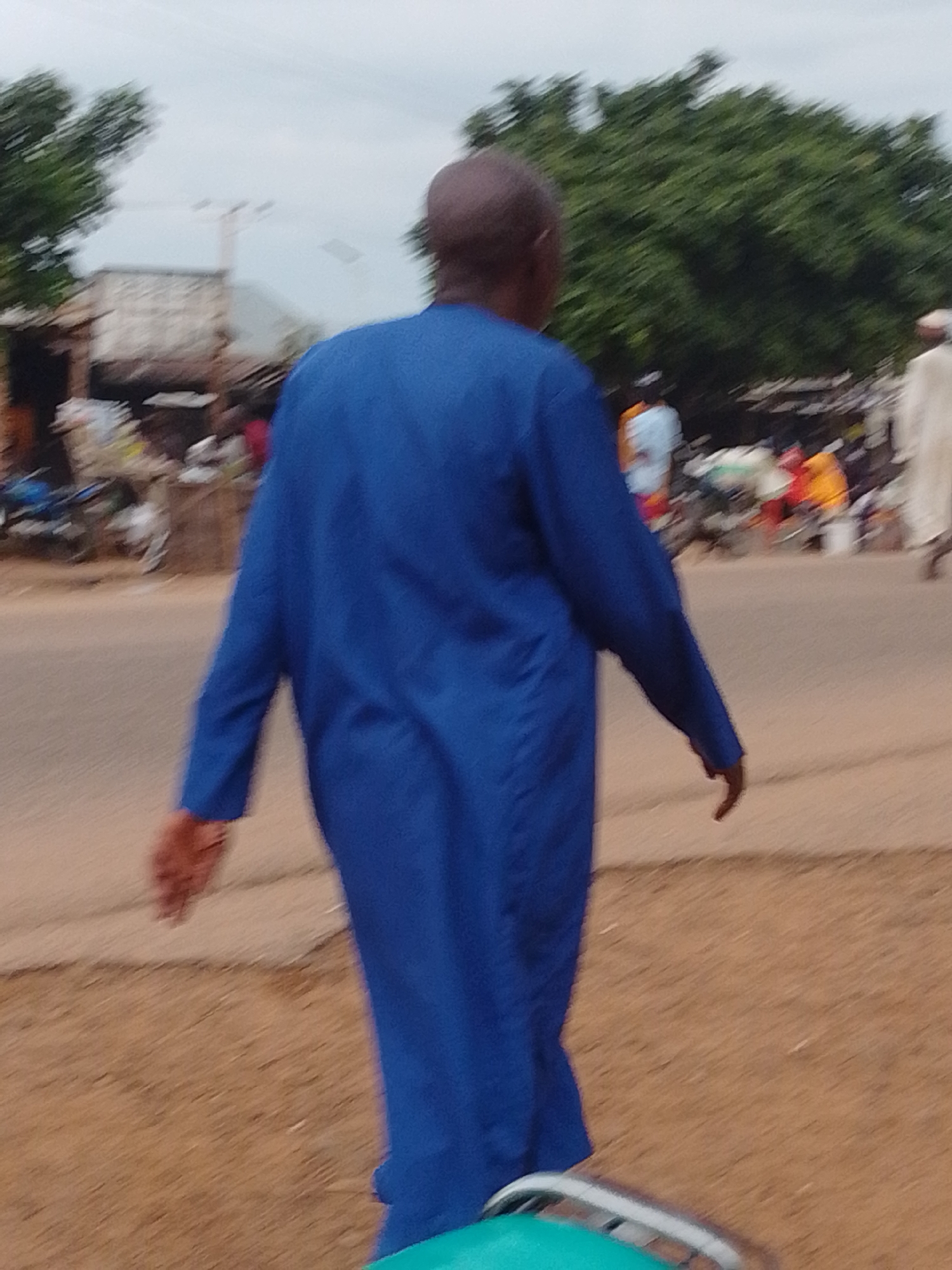 A person in a bright blue robe walks away from the camera on a dirt path next to a paved road in Gombe, Nigeria. The person has dark skin and is bald. In the background, there are several people in the distance, some wearing bright clothing, and a few motorcycles are parked. The environment appears to be a marketplace or a busy street with some buildings and trees visible. The sky is overcast.