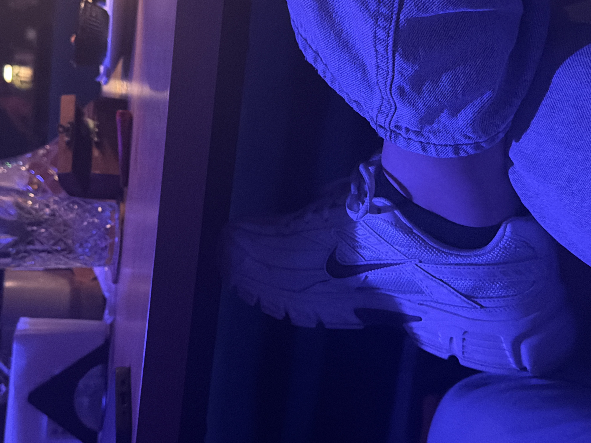 The image, oriented vertically, captures a close-up of a person's lower leg and foot within a dimly lit indoor setting in Plovdiv, Bulgaria. The scene is bathed in a strong blue and purple ambient light, suggesting an evening or nighttime atmosphere.
The subject is wearing blue denim jeans, visible from the mid-calf to just above the ankle, with the fabric showing texture and folds. On their foot is a white sneaker, clearly showing a black Nike swoosh logo on its side. The foot appears to be resting on a dark, indistinct surface or another object.
To the left of the person, a wooden table or counter surface is visible, laden with various objects. These include a glass or container filled with crumpled, reflective material (possibly foil or decorative elements) and indistinct bottles or glasses. Further down the table, a white rectangular object, perhaps a napkin holder or small box, is also present. Blurred lights in the background hint at a larger venue, possibly a bar, club, or restaurant, typical of a social gathering spot.
No direct activity is being performed other than the apparent resting of the foot. The notable details are the striking blue-purple lighting that colors the entire scene and the clear branding of the Nike sneaker. No other legible text is visible in the frame. The image provides an intimate, mood-driven snapshot of an individual's presence in a vibrant nightlife setting.