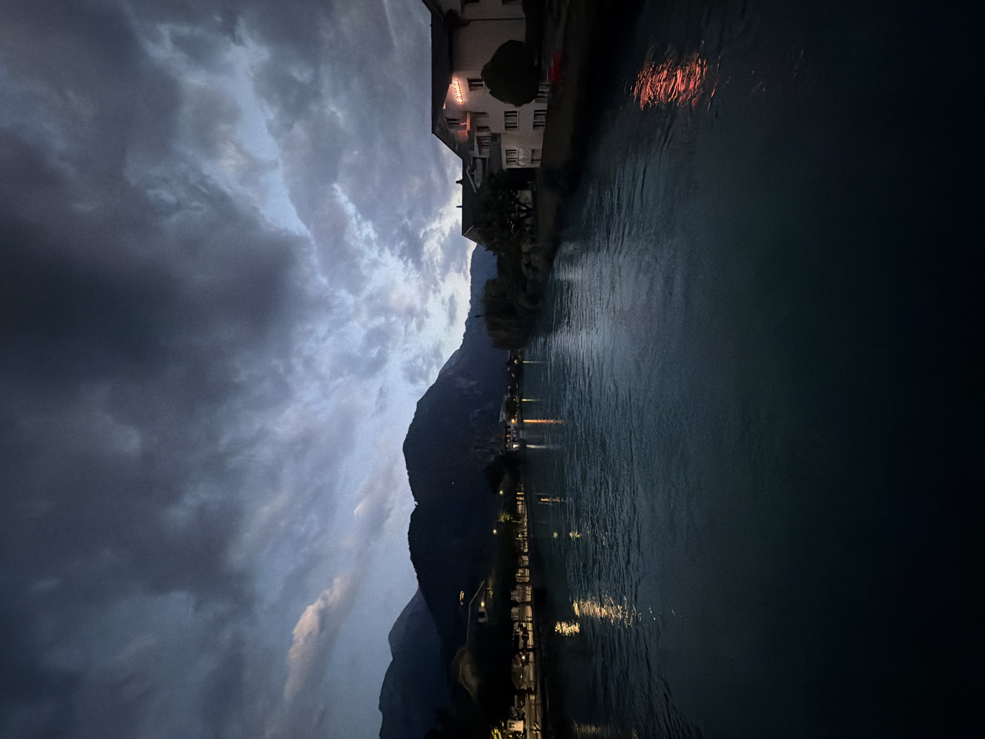 This is a nighttime photograph of the Aare River in Interlaken, Switzerland. The sky is dark and overcast with dramatic clouds. Mountains are visible in the background on the left side of the image, silhouetted against the sky. On the right bank of the river, there is a white building with red neon lettering that reads "Le Grand". The building is illuminated, and its reflection can be seen in the water. The river itself is dark and reflects the lights from the town. The scene is peaceful and serene, conveying a sense of calm despite the overcast sky.