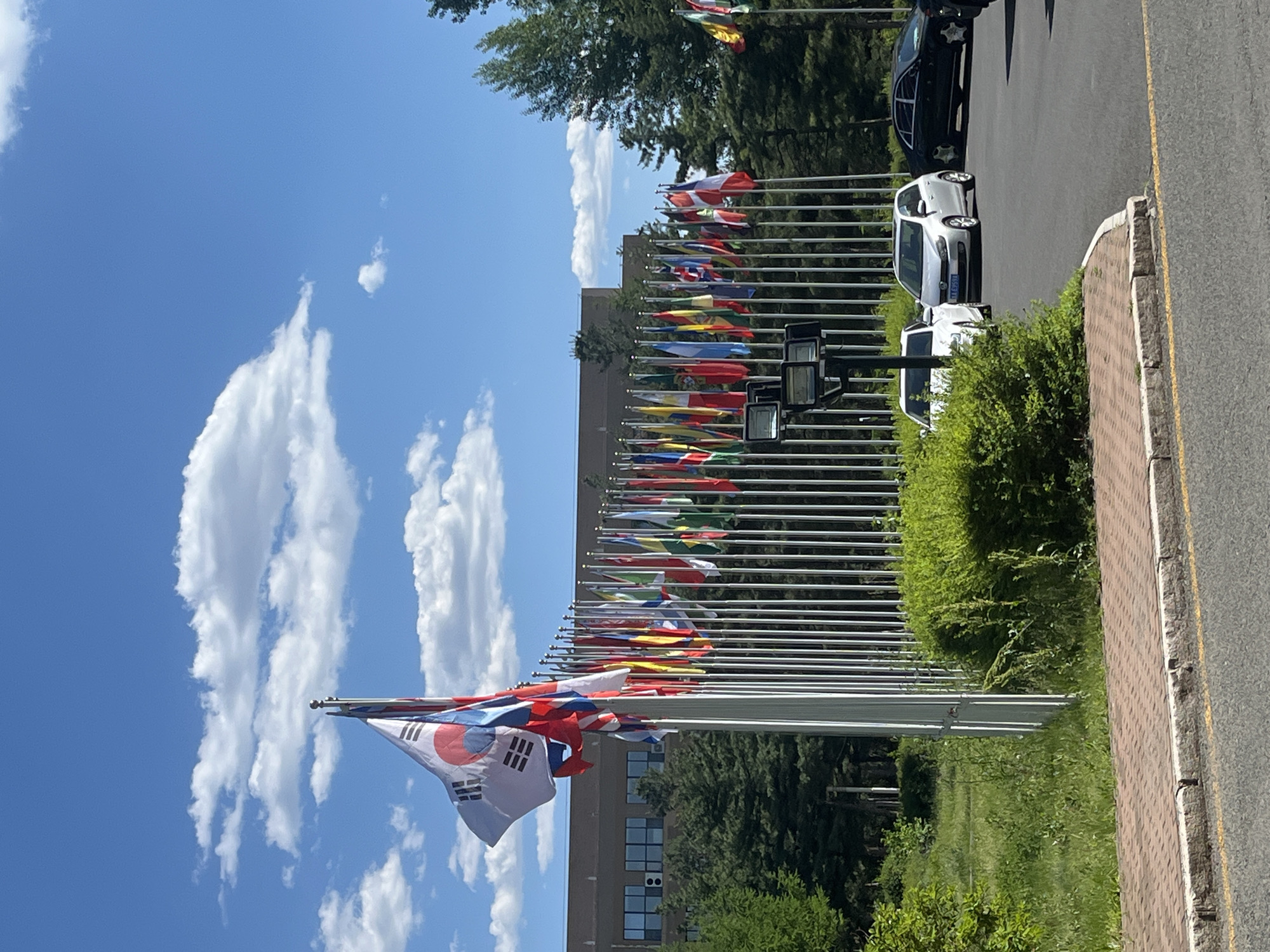 The image captures a scene in Changchun City, China, featuring a row of flagpoles displaying various national flags. The flags line a paved area alongside a road with parked vehicles. A building with visible windows and air conditioning units is situated behind the flagpoles, partially obscured by trees. The sky is blue with scattered white clouds. The South Korean flag is prominently displayed in the foreground. The overall environment appears to be outdoors during daylight hours.