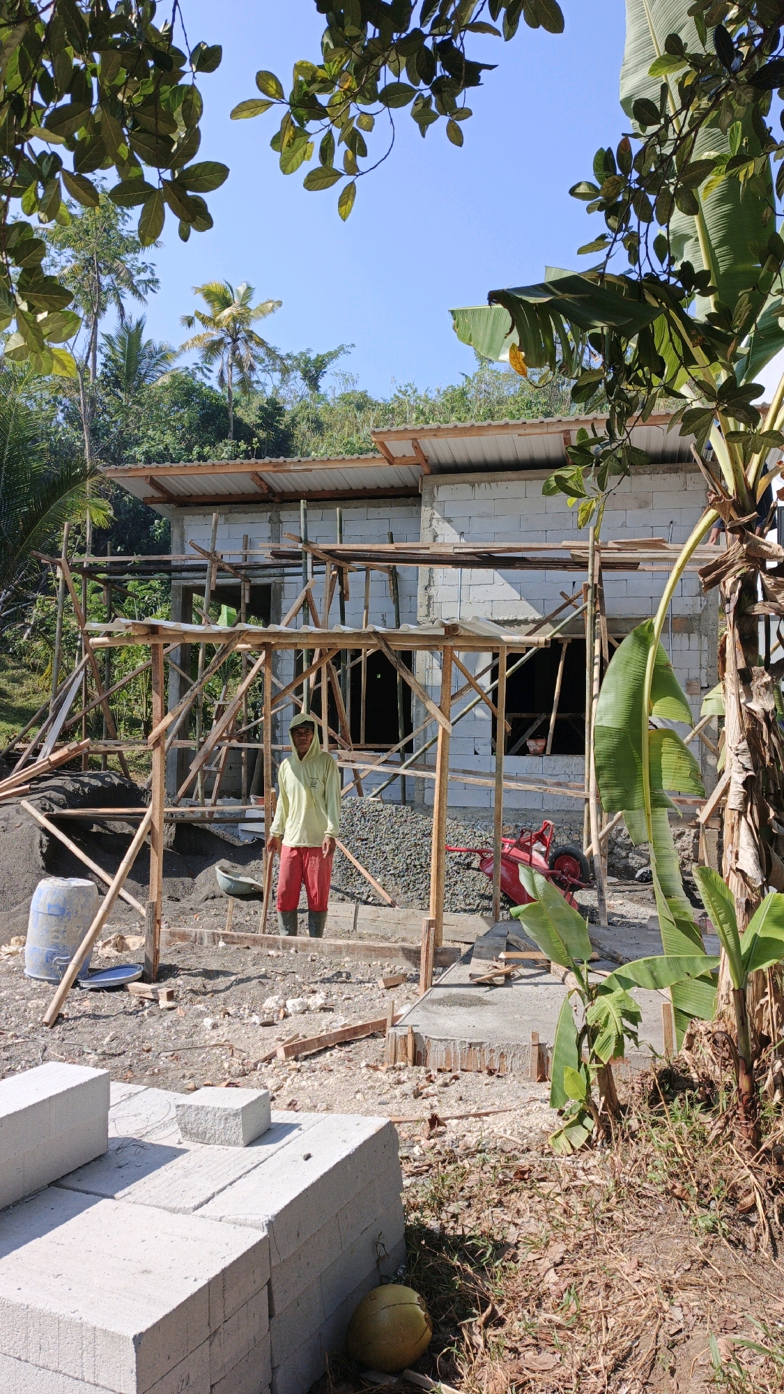 A full shot captures a construction site in Desa Batukaras, Indonesia, under a clear blue sky. In the foreground, stacks of white concrete blocks are neatly arranged on the dusty ground. A lone coconut rests near the blocks. A man, wearing a light yellow hooded shirt and red trousers, stands in the middle of the construction area, holding what appears to be a shovel. He is positioned within a wooden scaffolding structure. Behind him, a partially constructed building, made of white cinder blocks, has a corrugated metal roof. The surrounding environment is lush with tropical vegetation, including palm trees and banana trees, indicating a rural or semi-rural setting. Construction materials, such as sand and gravel, are visible scattered across the ground, along with a red wheelbarrow. The scene suggests ongoing construction work in a tropical climate.