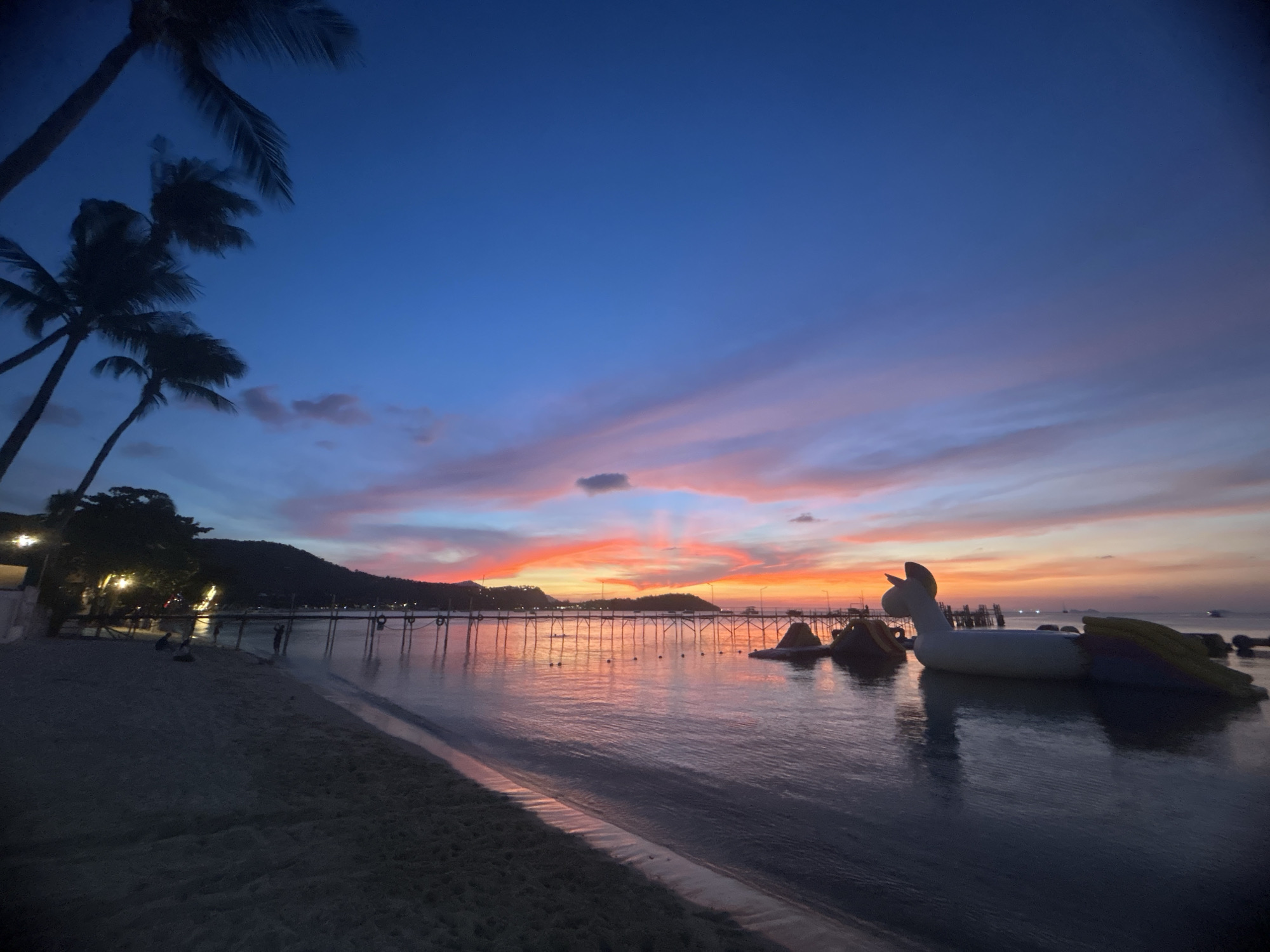 The media file captures a picturesque tropical sunset at Baan Bang Rak, Thailand. The scene is dominated by a vibrant sky, transitioning from a deep indigo blue at the top to brilliant oranges, reds, and pinks near the horizon, with some soft, colorful clouds scattered across.
In the foreground on the left, a sandy beach curves gently, appearing dark in the twilight. Silhouetted palm trees frame the upper left corner, their fronds catching the last light. Along the shoreline, hints of artificial lights from establishments are visible, and a few indistinct figures can be seen on the beach.
The center and right foreground are filled with the calm water, which beautifully reflects the rich hues of the sunset sky. A large, white inflatable unicorn or similar fantasy creature floats prominently on the water, suggesting a recreational area or water park. Other smaller inflatables are visible nearby.
Stretching across the mid-distance is a long wooden pier, illuminated by string lights that cast shimmering reflections on the water. Distant mountains or hills are silhouetted against the glowing horizon, adding depth to the panoramic view.
The overall atmosphere is serene and tranquil, capturing the end of a day in a tropical paradise. The time of day is clearly sunset, as indicated by the dramatic sky colors and fading light. There is no visible text in the image.